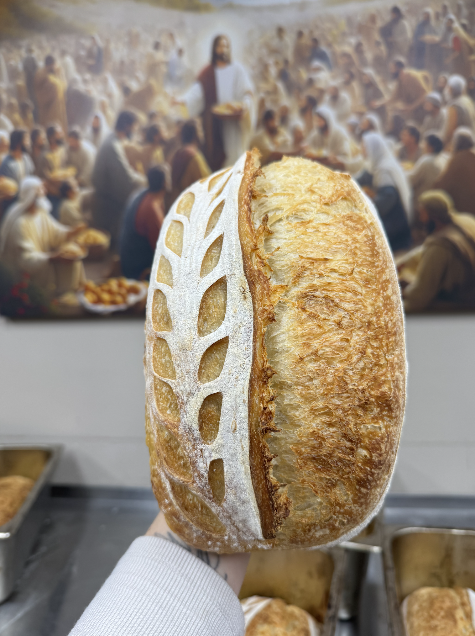 A large piece of toasted sourdough bread with a crispy crust and a lattice of white flour on the surface, held in a person's hand with a background of a religious gathering with people sitting and Jesus figure in the center.