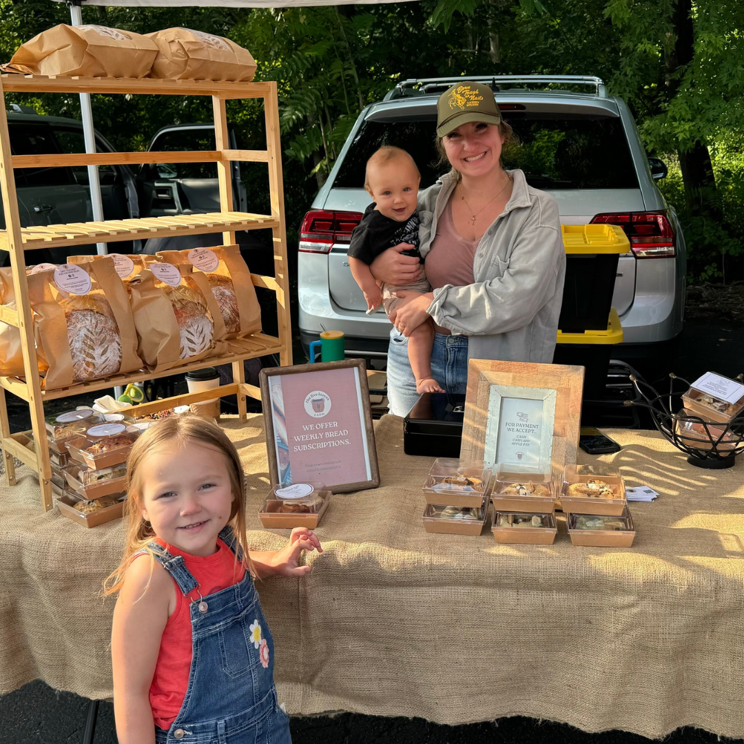 A holding a smiling toddler at a bakery stand outdoors, with a young girl in a red shirt and denim overalls in front. The stand has bread and baked goods on tables and shelves, with signs advertising weekly bread subscriptions and a cake for payment.