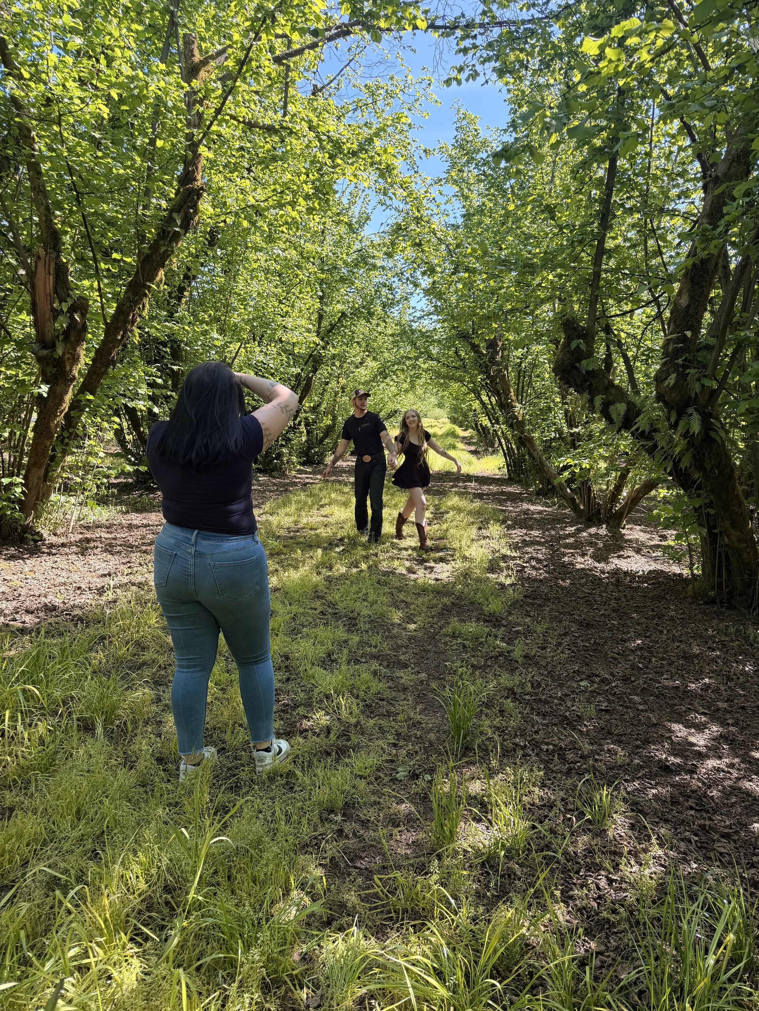 A woman with tattoos on her arms takes a photo of a man and a woman walking hand in hand through a green, sunny forested path surrounded by trees with bright green leaves.