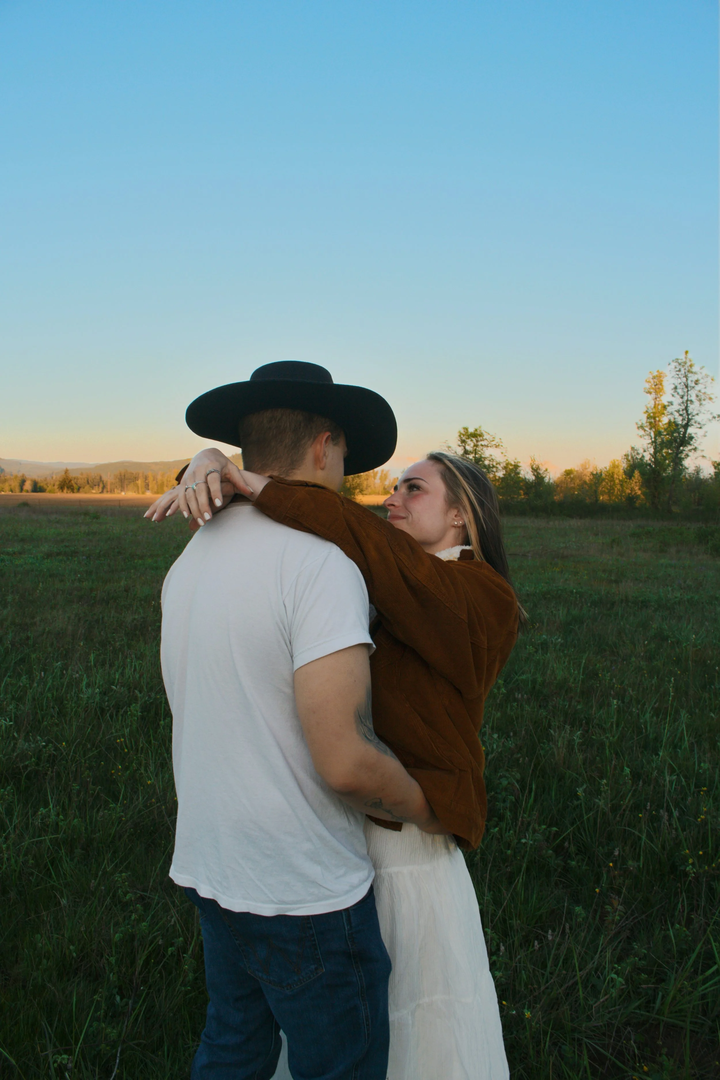 A couple hugging in a field during sunset, with trees and mountains in the background.