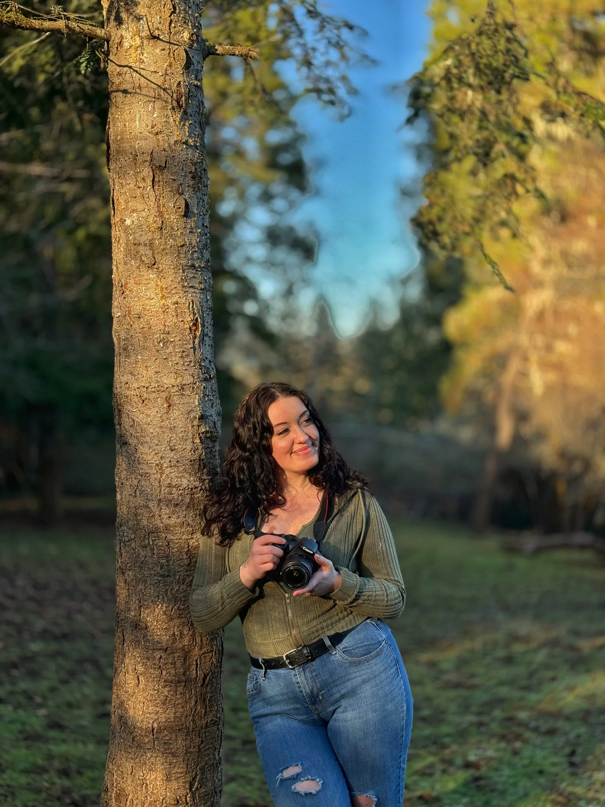A woman with curly dark hair smiling and holding a camera, leaning against a tree in a park during daylight, with a background of green trees and blue sky.