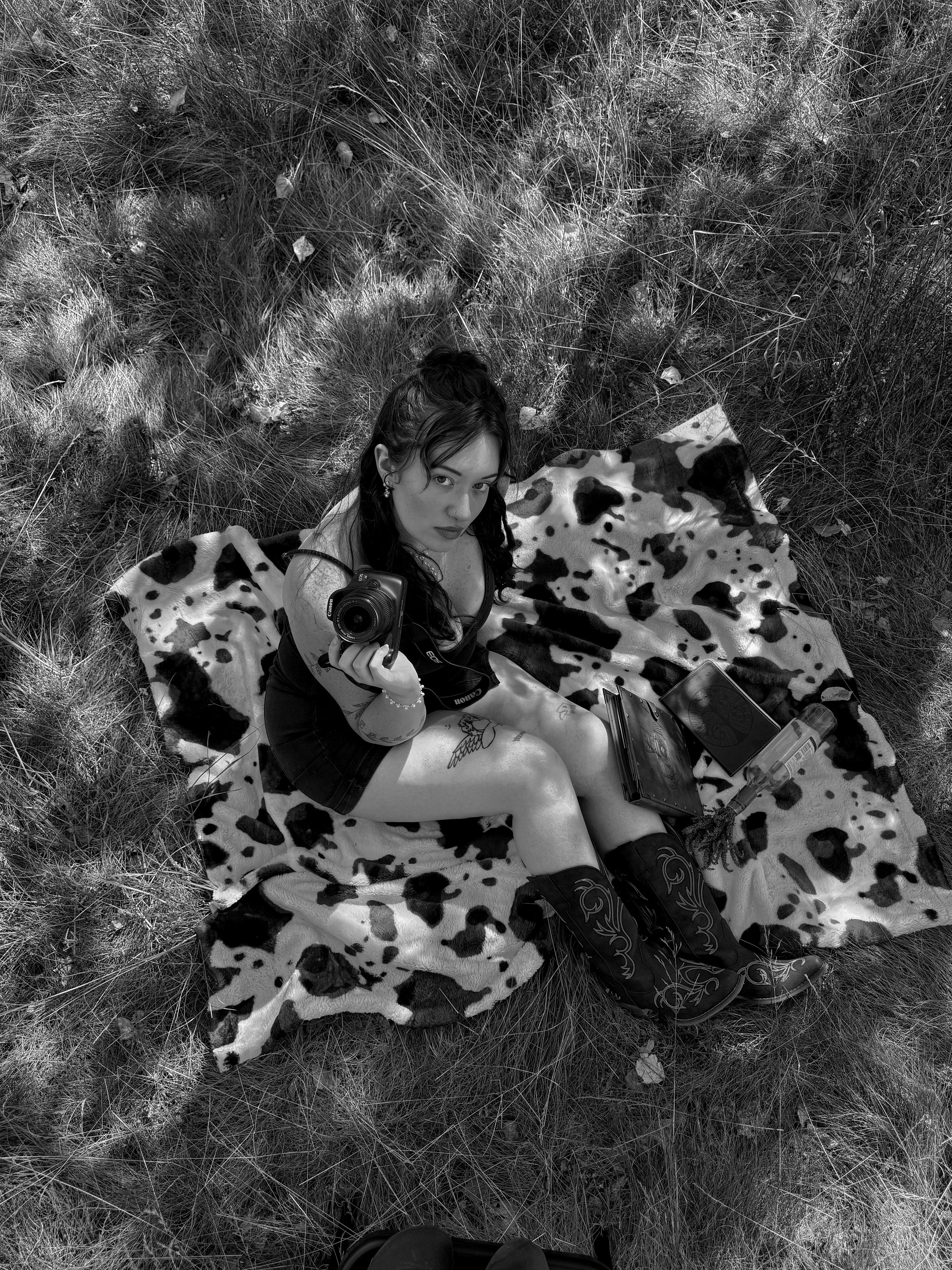 A woman with long dark hair and tattoos on her arm, sitting on a cow-print blanket in a grassy outdoor area, taking a selfie with a camera. There are various objects around her, including a laptop and a water bottle.