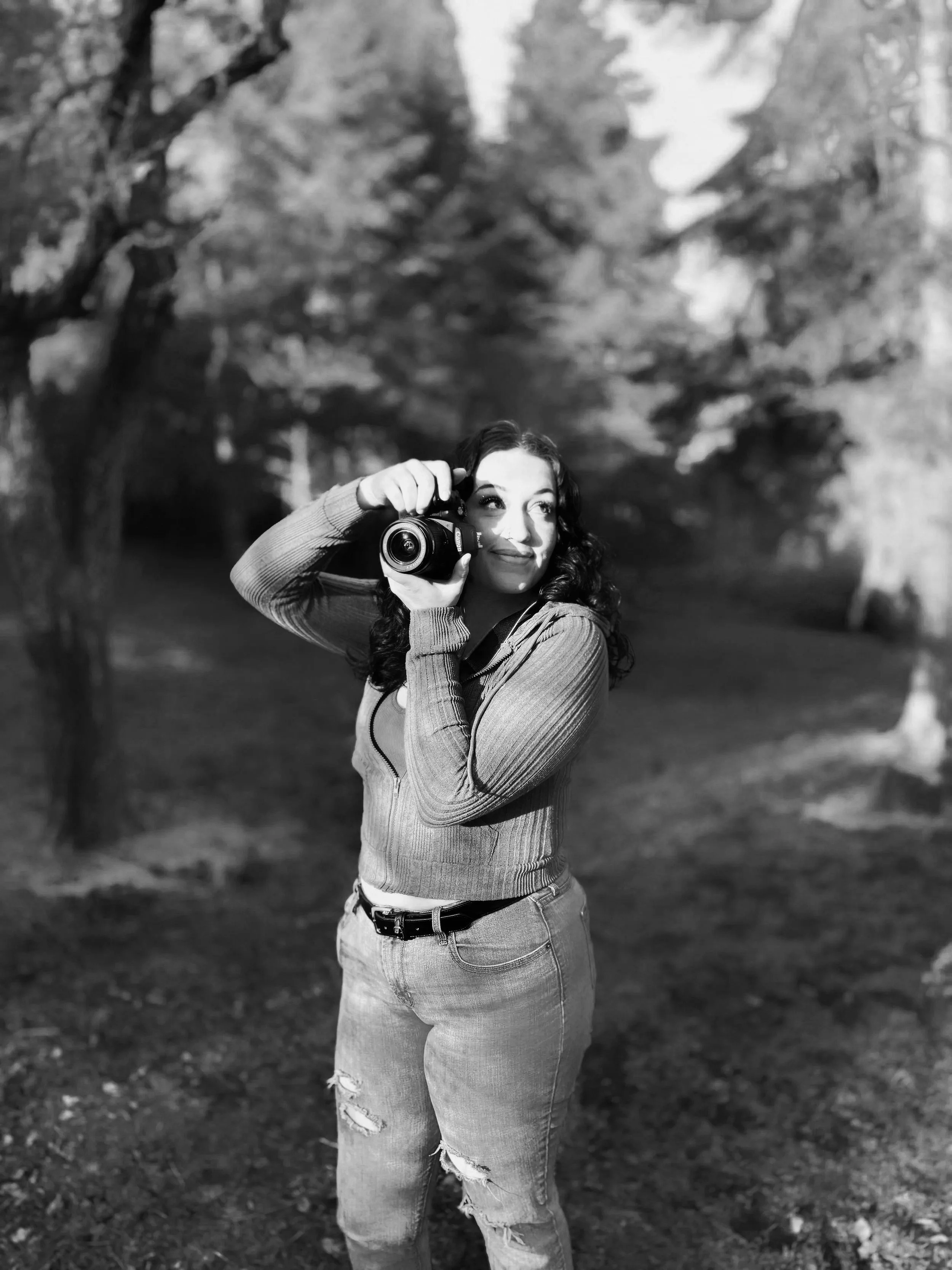 A woman with curly hair aiming a camera outdoors on a sunny day, background of trees and grass in black and white.