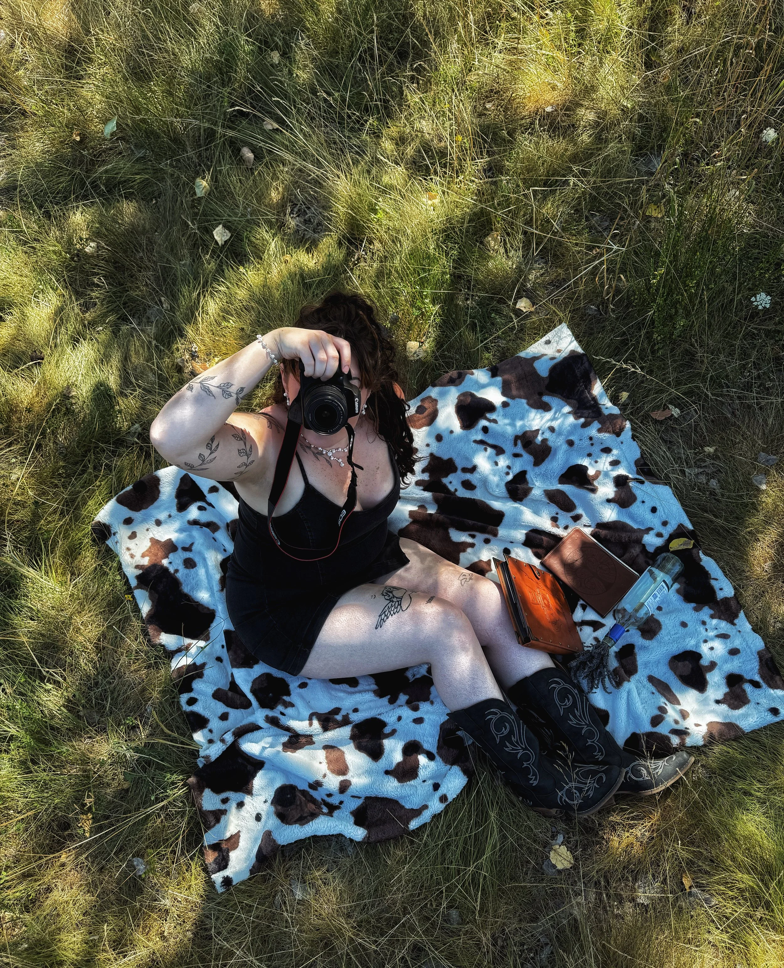 Person lying on a cow-print blanket outdoors in tall grass, taking a photograph with a camera. Wearing a black dress, boots, and jewelry, with various personal items nearby.