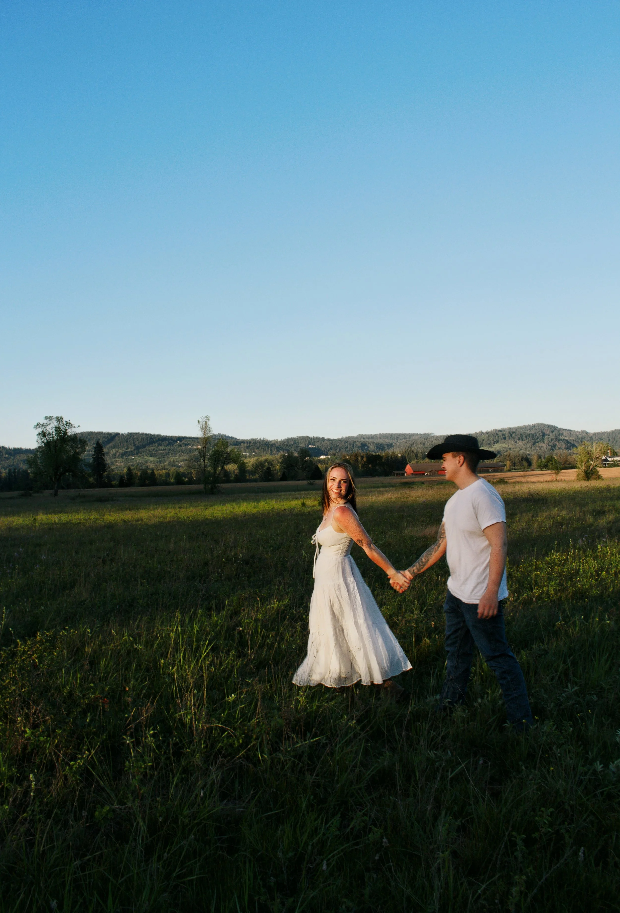 A smiling woman in a white dress holding hands with a man wearing a cowboy hat and white T-shirt in a green field with mountains in the background.