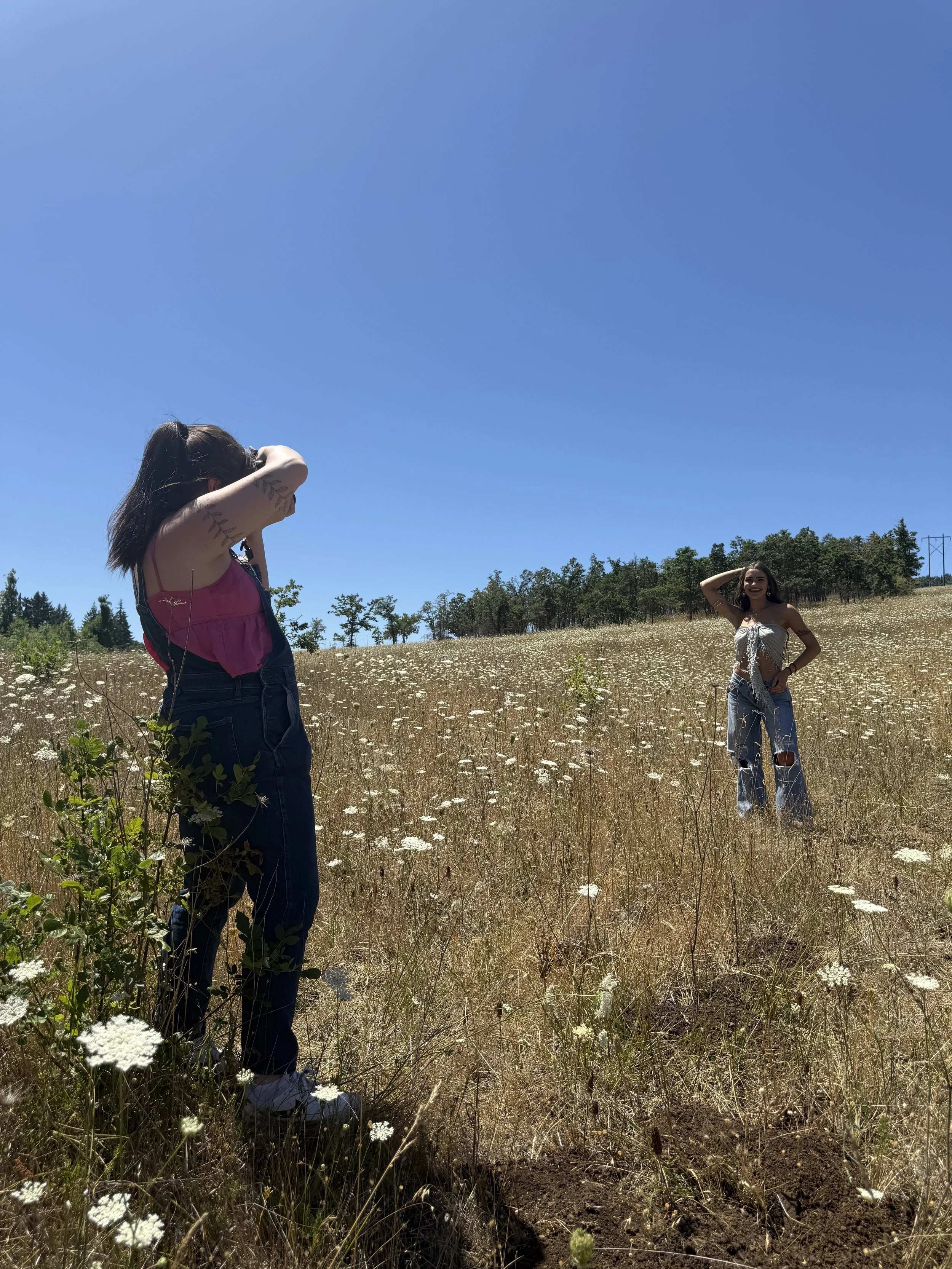 Two women standing in a meadow with white flowers under a clear blue sky, one adjusting her hair and the other smiling.