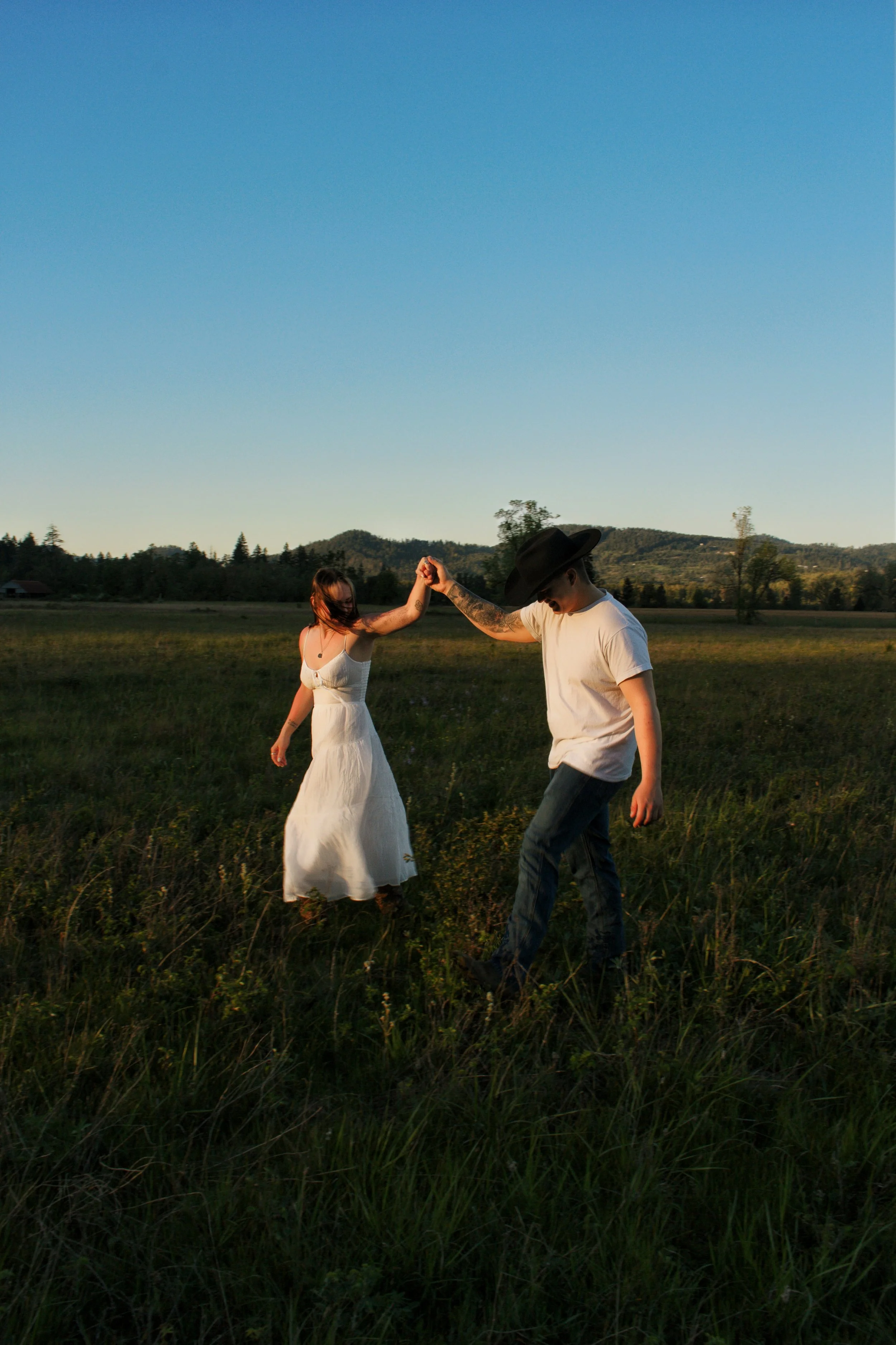 A man and a woman dancing hand in hand in a grassy field during sunset with mountains and trees in the background.