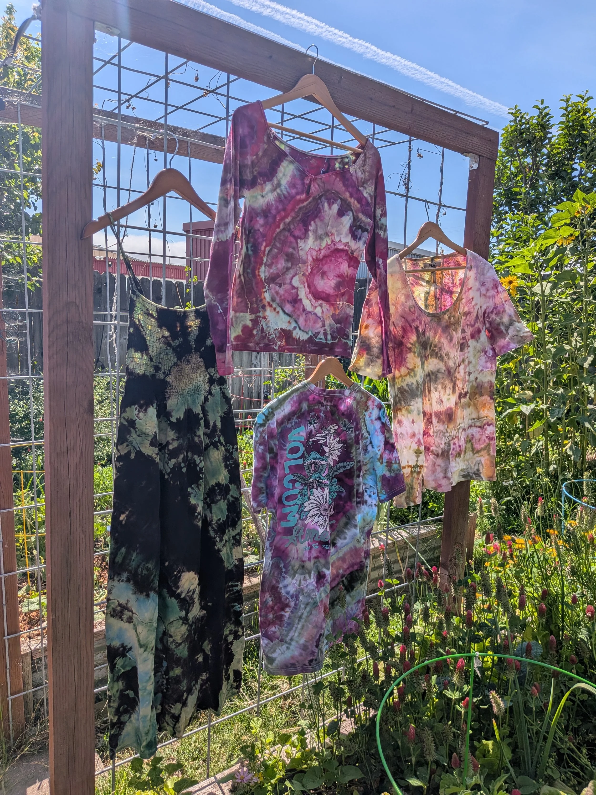 Colorful tie-dye shirts hanging on hangers on a wooden and wire outdoor clothing rack in a garden with flowers and greenery.