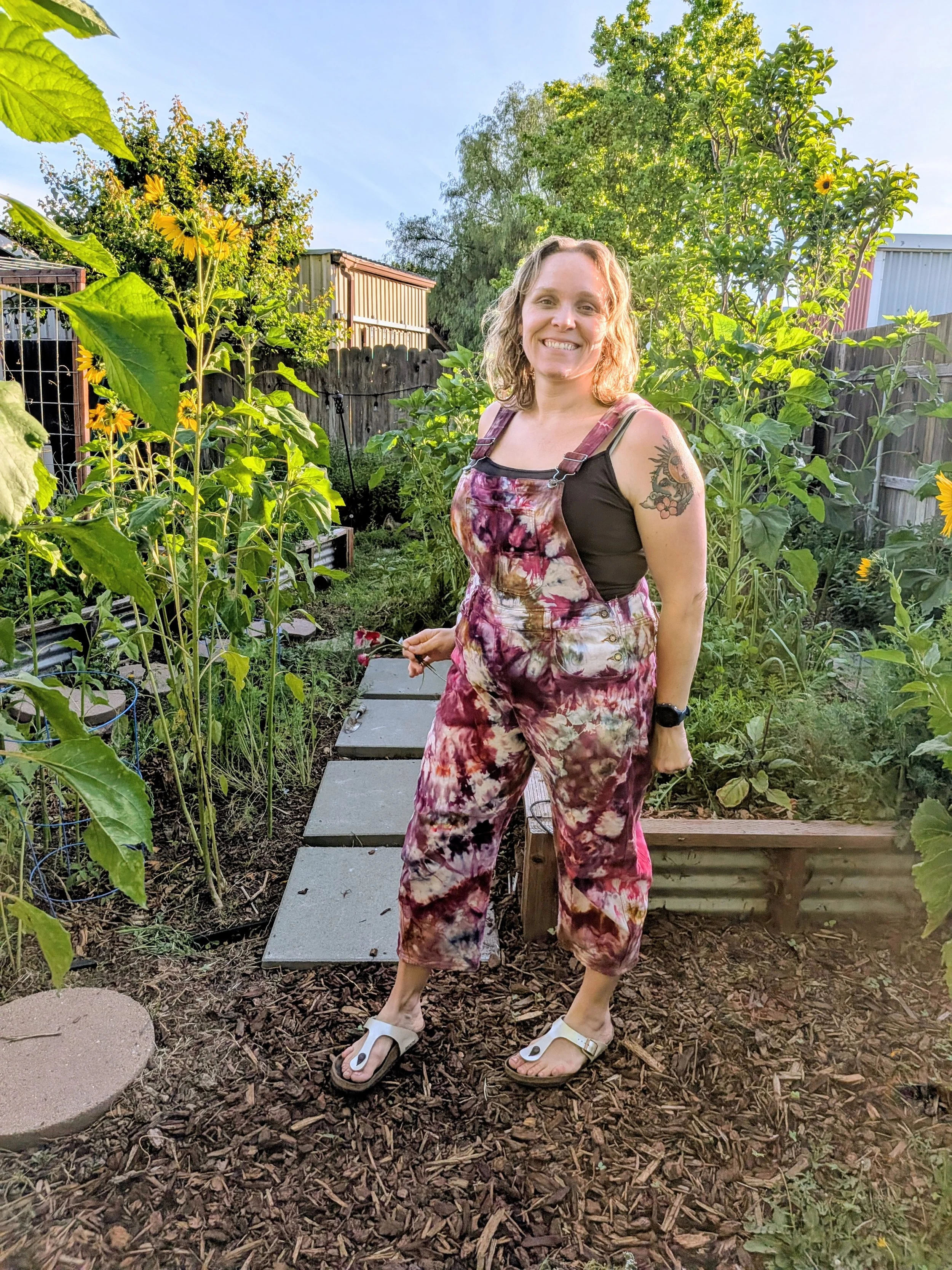 Shannon, the artist of Hazy Fibers standing on a garden path, surrounded by tall sunflowers, wearing and dyed ice-dye geode overalls and sandals, with a cheerful smile, in a backyard garden during daytime.