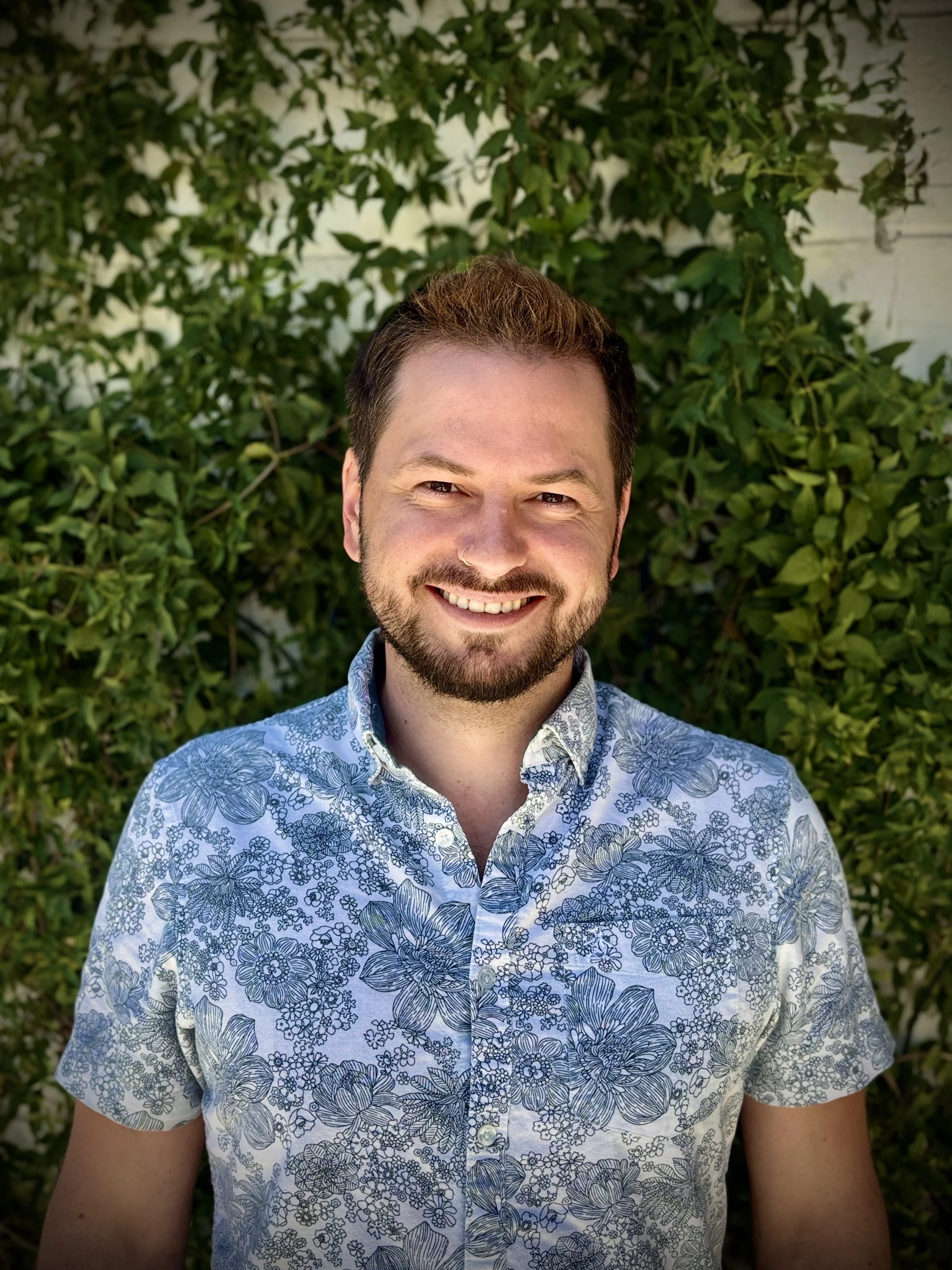 A smiling man with a beard and mustache, wearing a floral patterned shirt, standing outdoors in front of green leafy bushes.