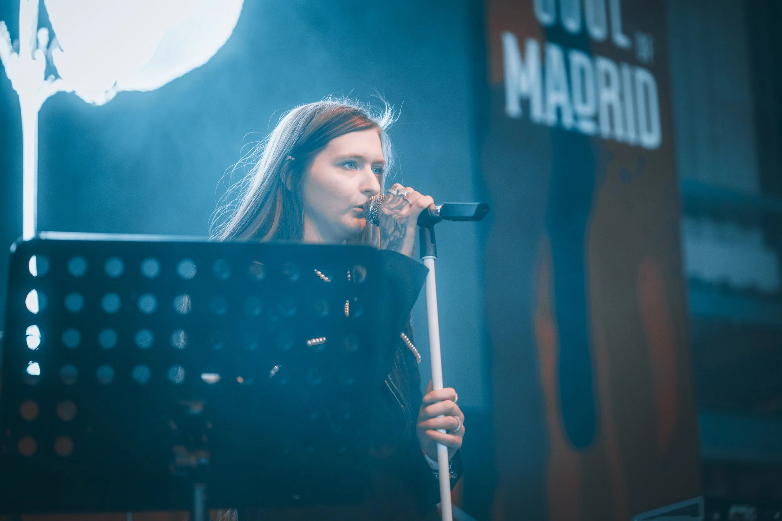 Woman singing into a microphone on stage with a music stand in front of her, blue lighting, and a blurred background with text.