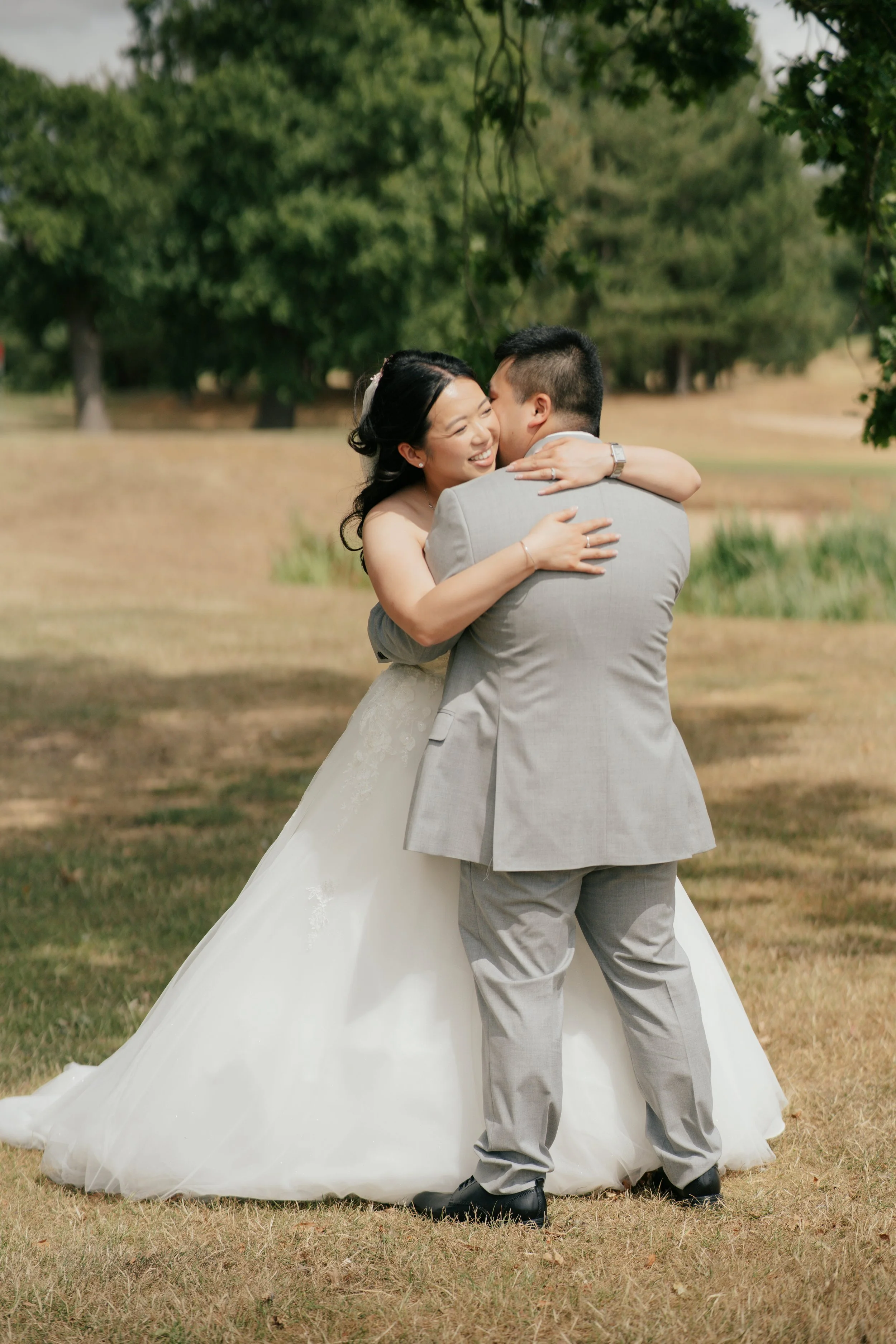 A bride and groom share a joyful embrace outdoors, with the bride smiling and the groom holding her, in a park with trees and grass.