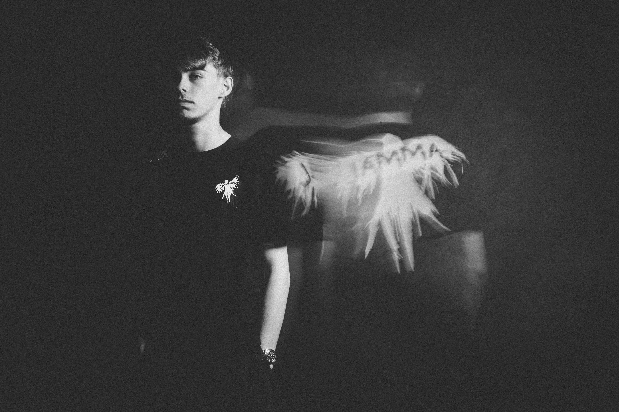 Black and white photo of a young man with short hair wearing a t-shirt, standing in a dark, minimalistic setting with a silhouette of a woman with long hair and a fringed top in the background.