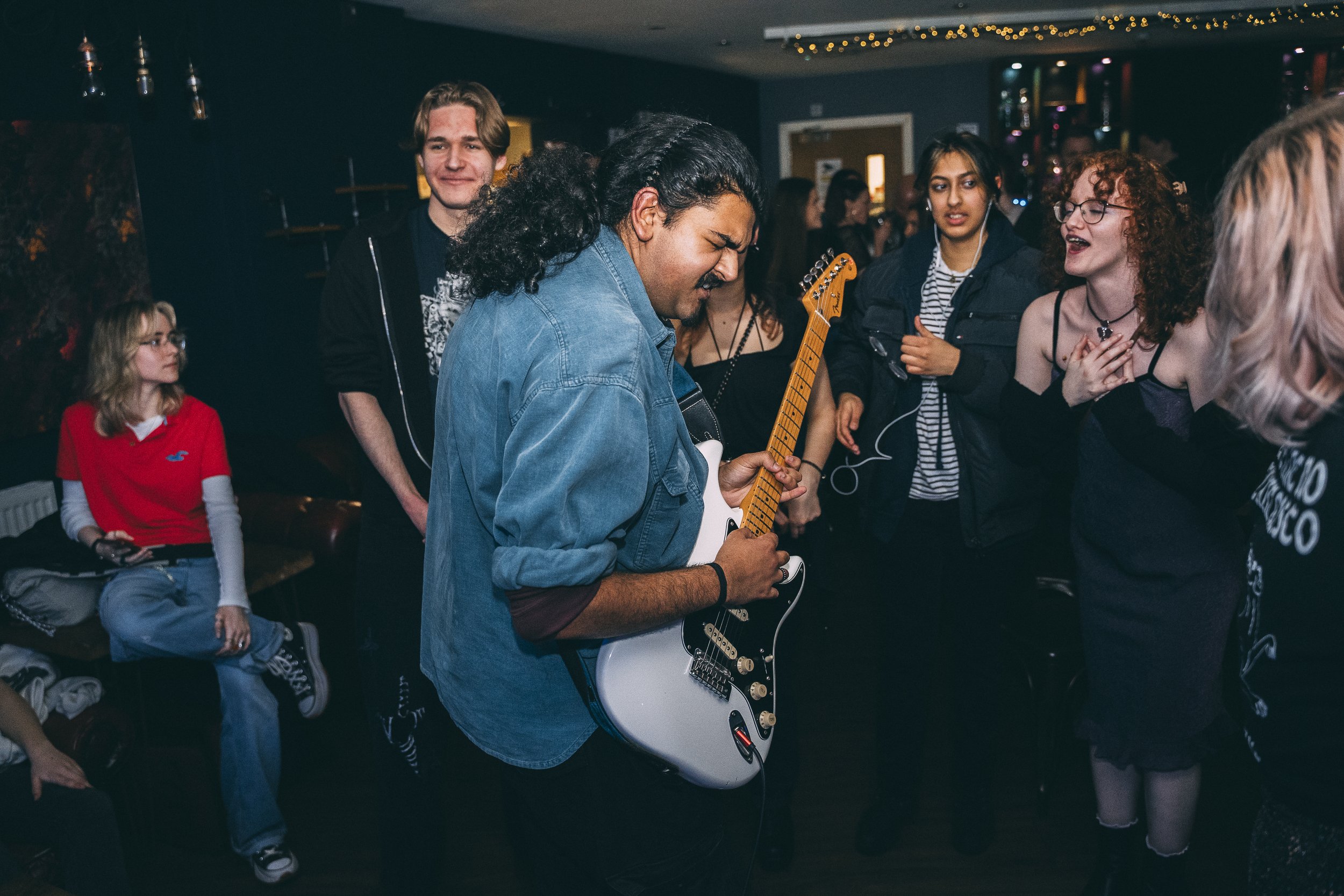 A group of young people, including a man with long, dark curly hair playing an electric guitar, in a dimly lit room decorated with string lights, enjoying music and socializing.