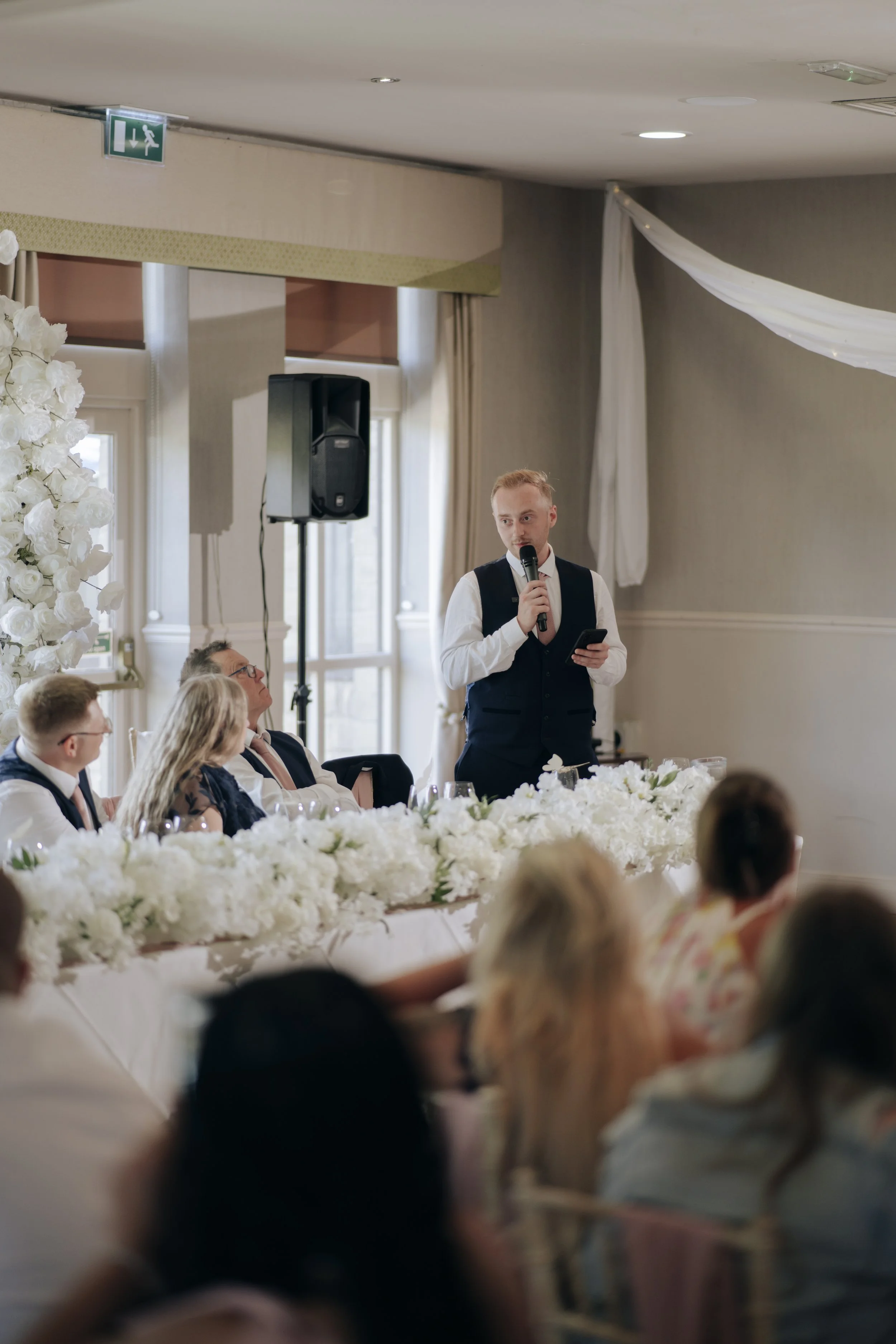 A man in a vest and tie holding a microphone speaks at a wedding reception, with guests seated at long tables decorated with white floral arrangements, in a bright indoor setting.