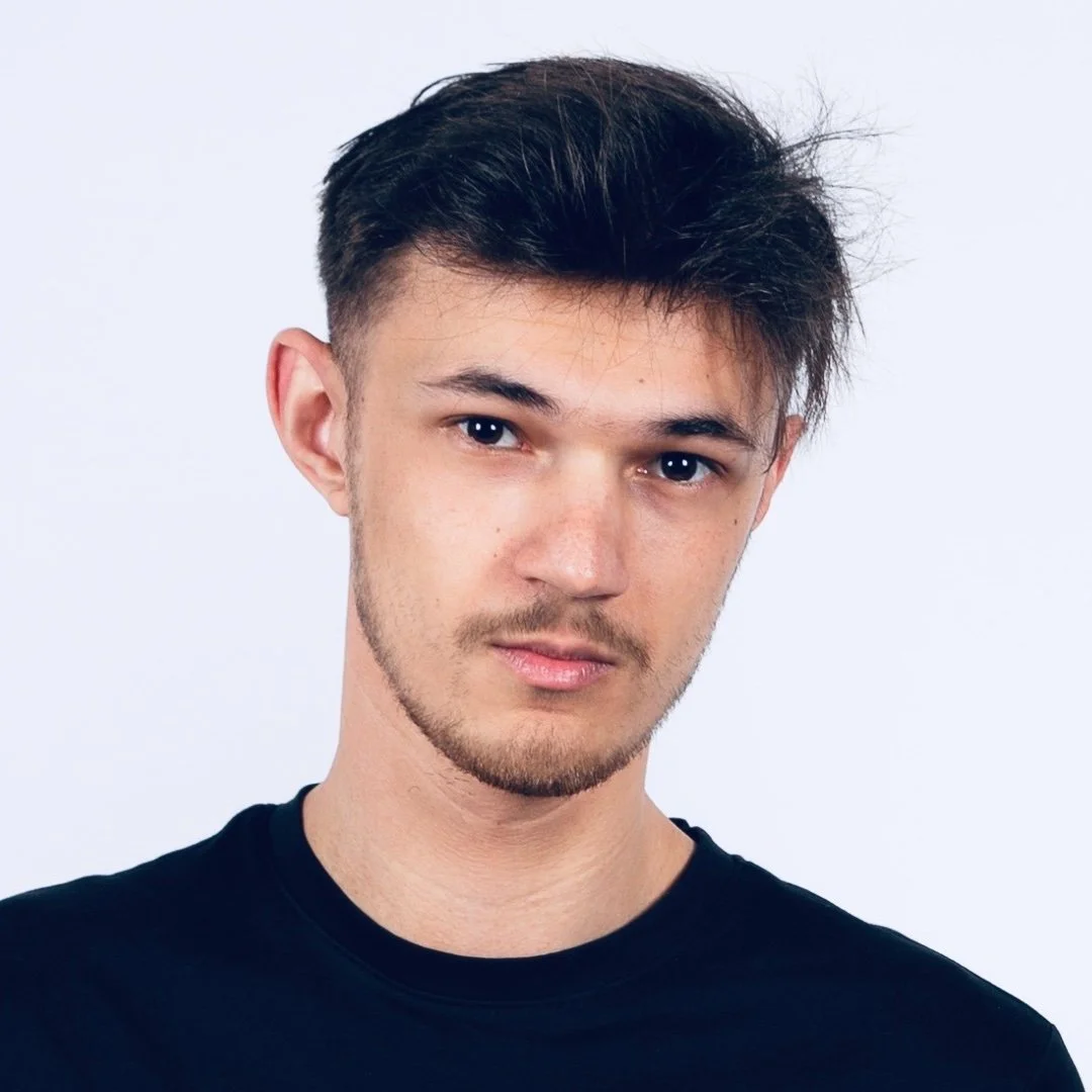 Close-up of a young man with short, dark hair, light facial hair, and wearing a black shirt, looking into the camera with a neutral expression.