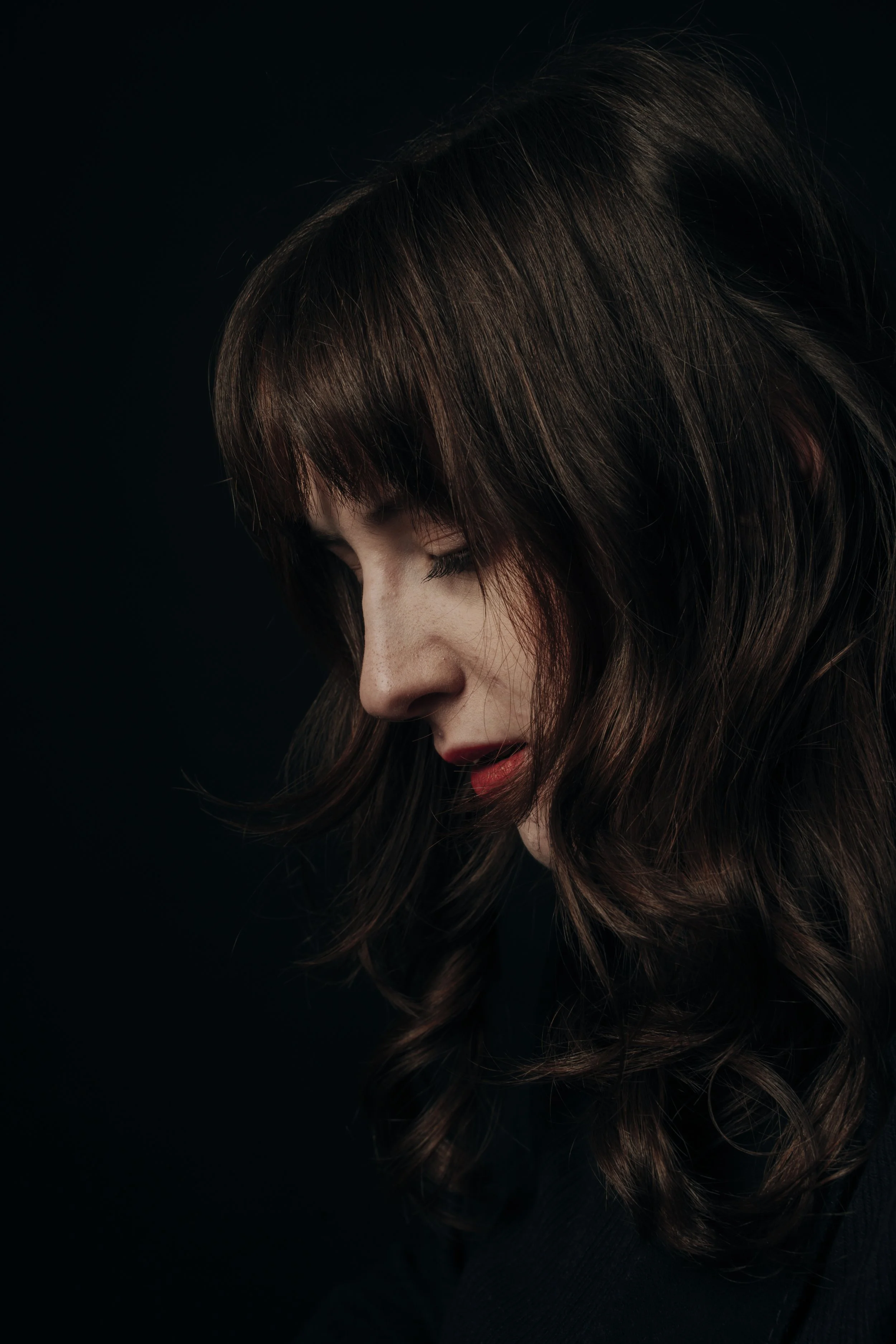 Close-up of a woman with brown hair and faint makeup, looking downward against a black background.