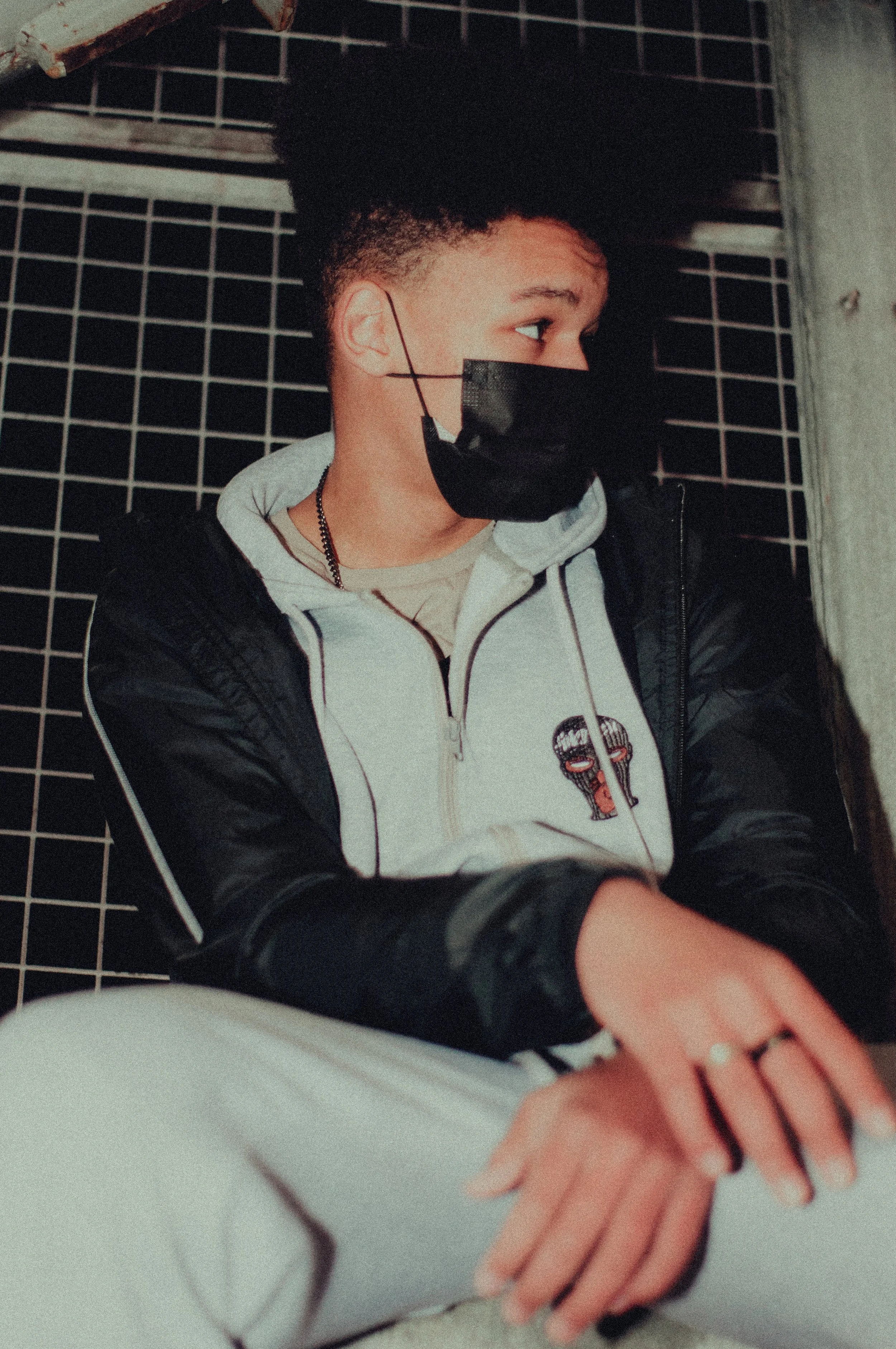 A person wearing a black face mask, a gray hoodie, and a black and gray jacket, sitting against a black grid wall, with their hair styled in a high curly afro, and looking to the side.