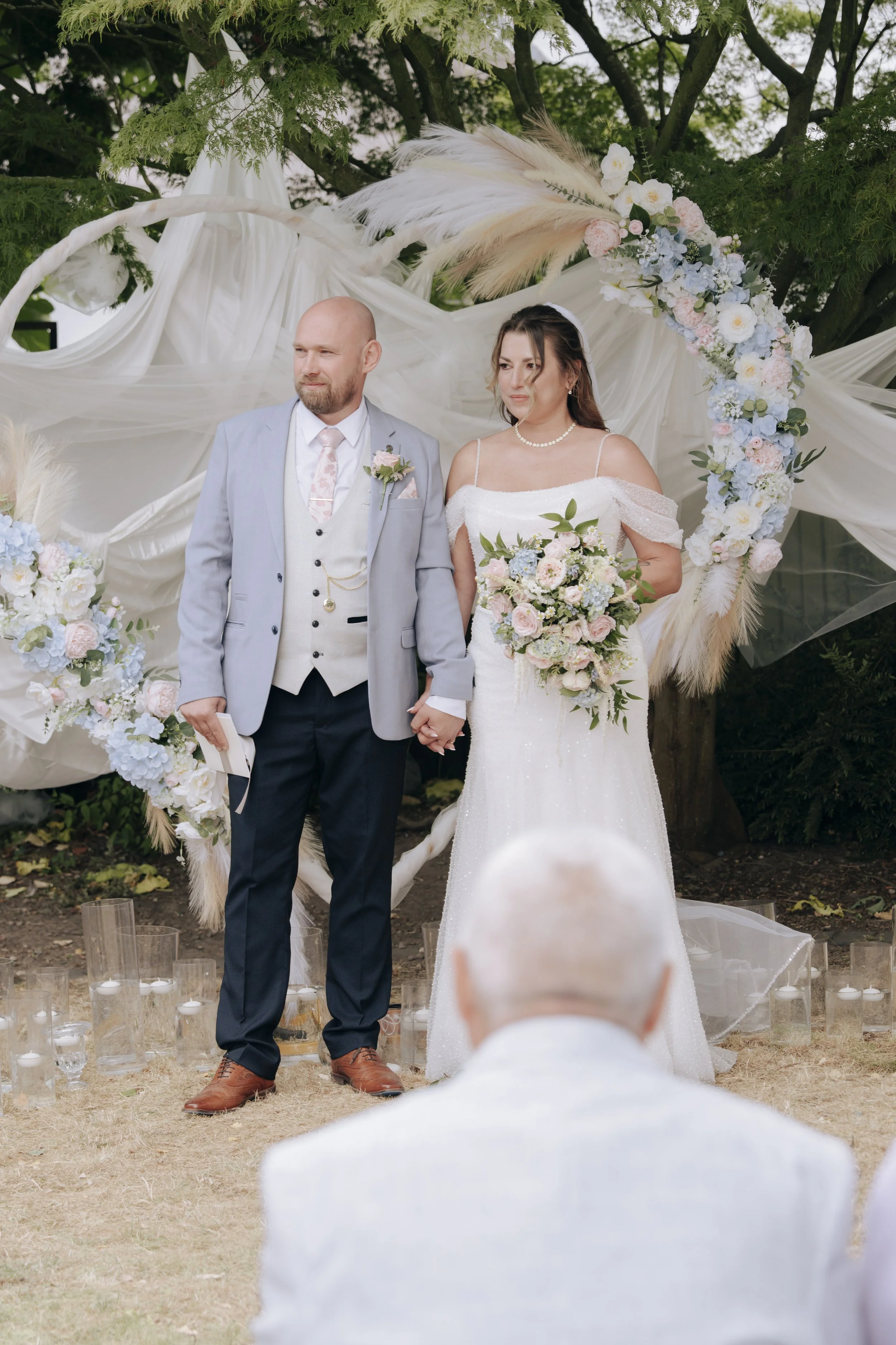 A bride and groom standing outdoors during their wedding ceremony, holding hands, with a decorative floral arch behind them, and a guest seen in the foreground.