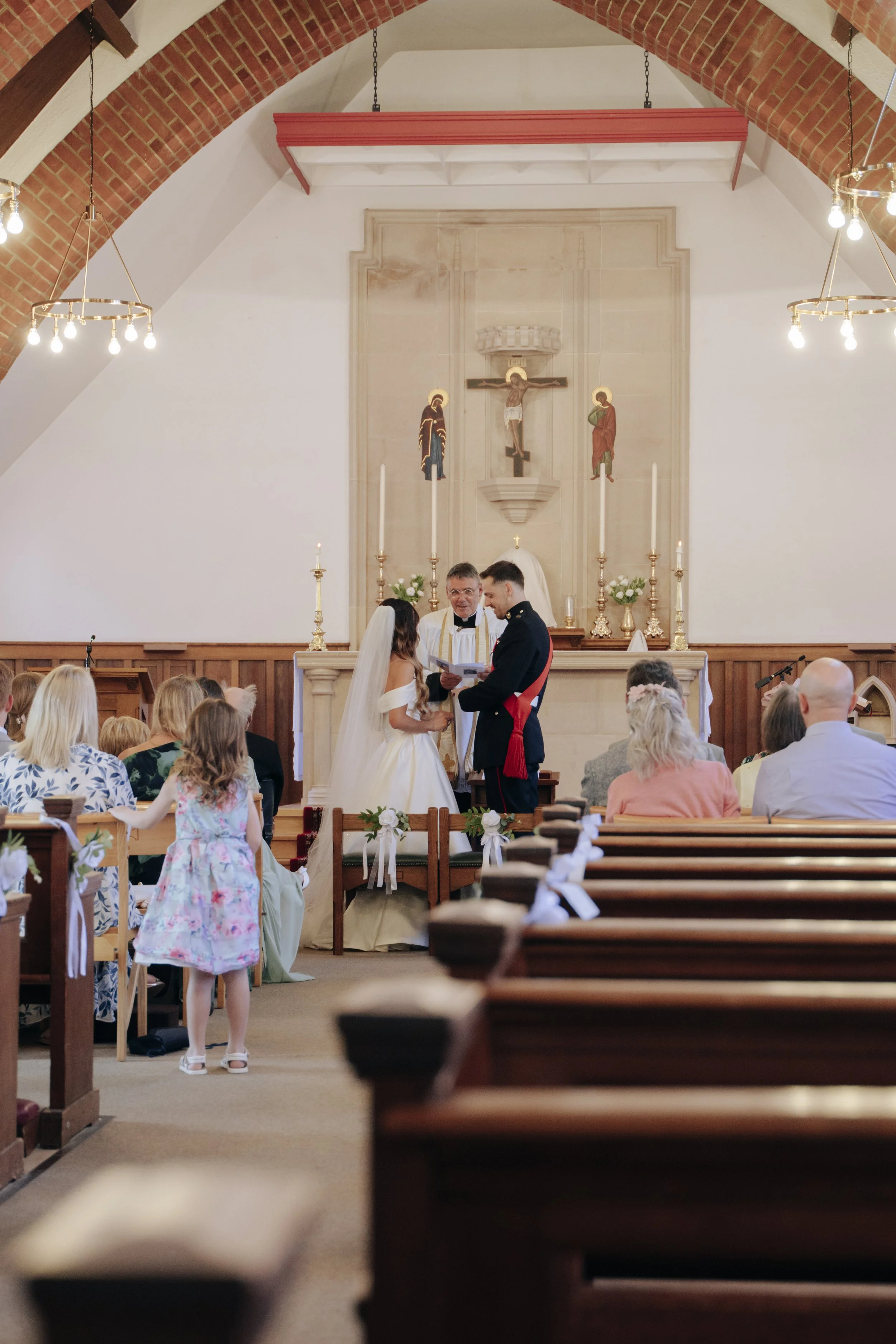 A wedding ceremony taking place inside a church with wooden pews, a bride and groom facing a minister, and guests seated on both sides. The church has a decorated altar with a crucifix and icons, and brick arch ceilings with hanging lights.