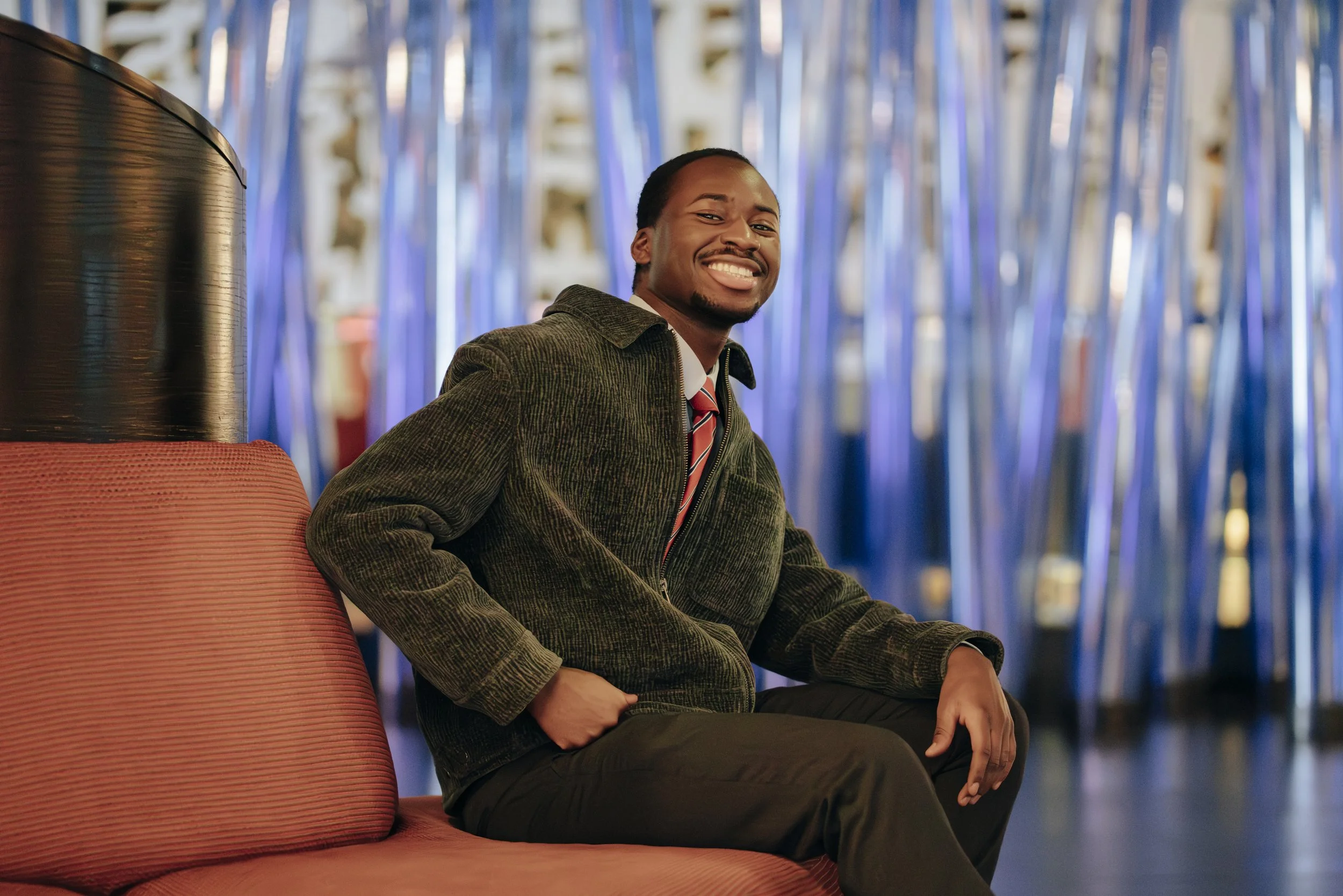 A smiling man with dark hair and a goatee, dressed in a blazer and striped tie, sitting on a pink couch in front of shiny blue and silver decorations.