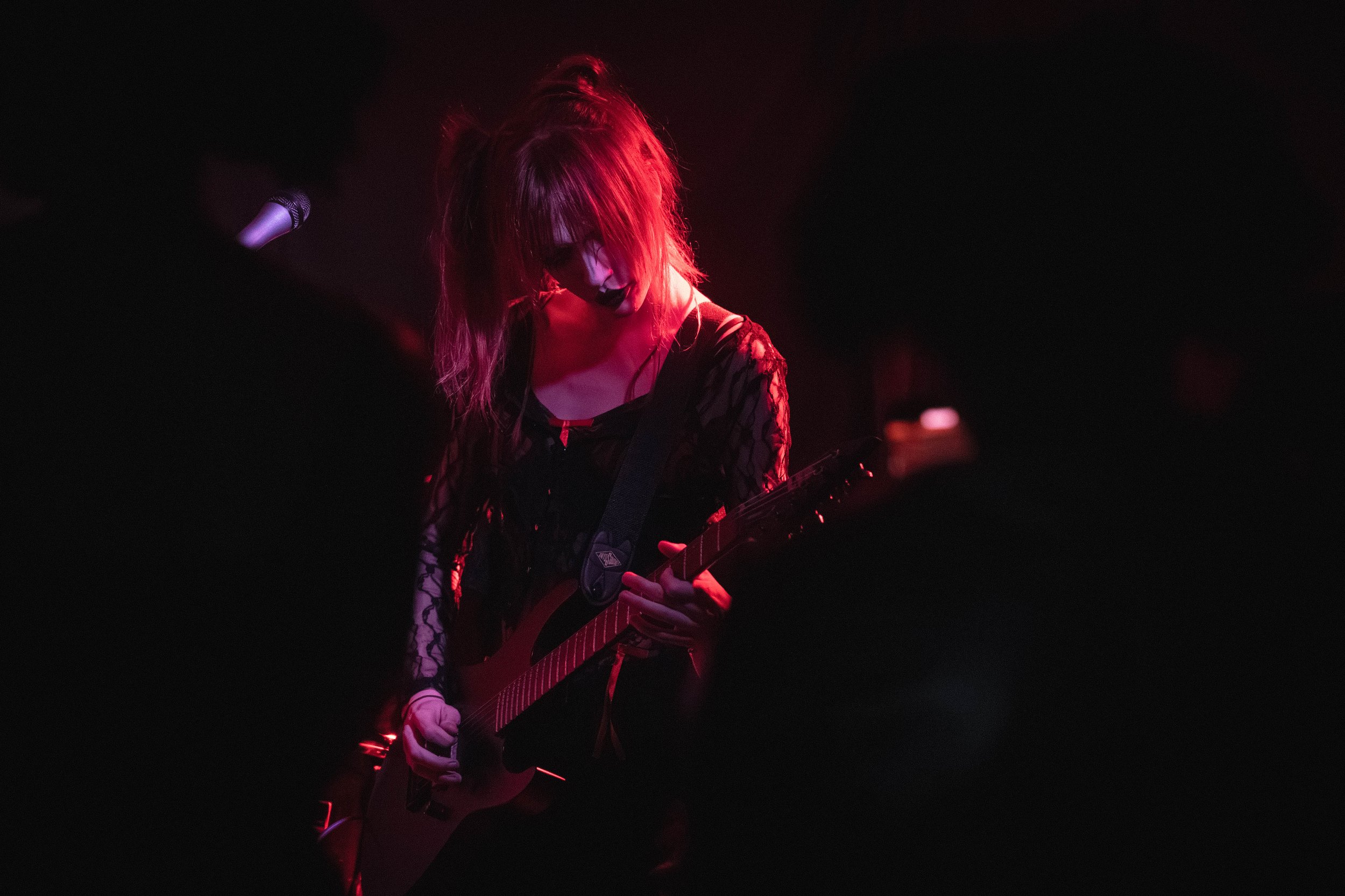 A female rock musician playing an electric guitar on stage, illuminated by red lighting, with a microphone stand nearby, dark background.
