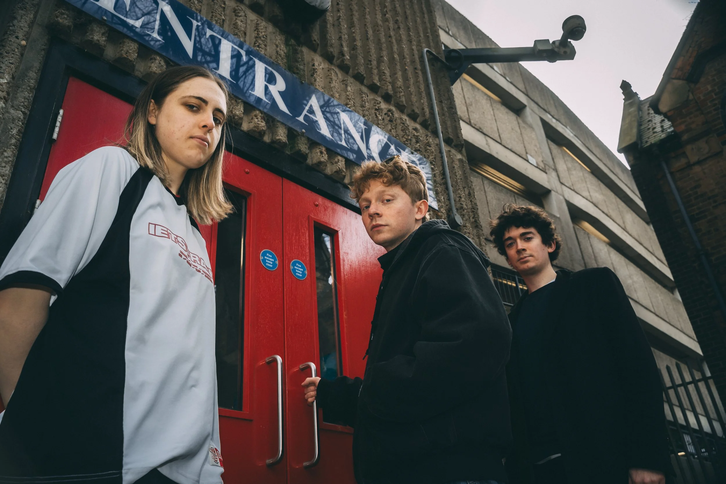 Three young people standing in front of a red door on a city street, with a blue street sign reading 'entrance' above them.