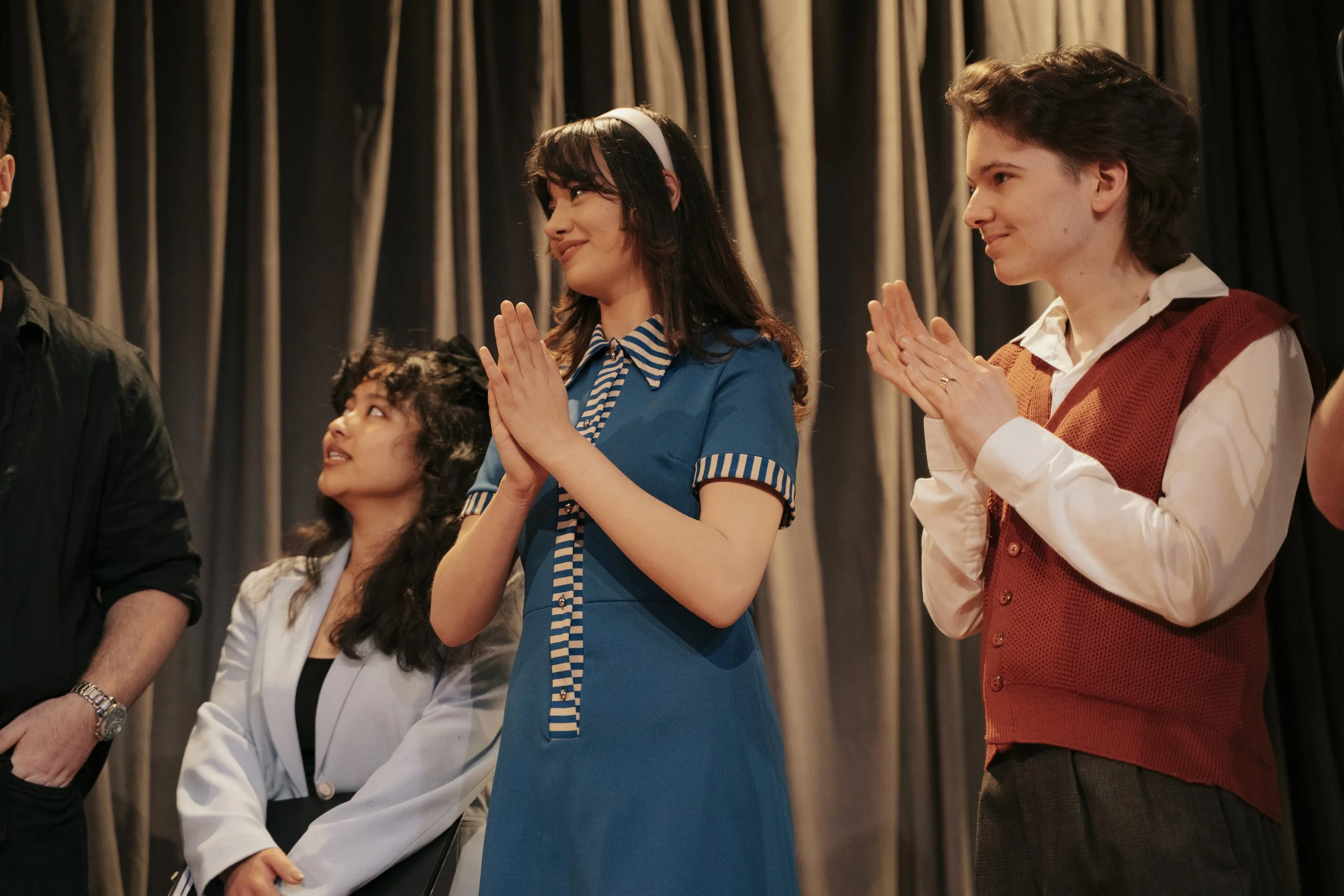 People on stage at an event, standing with hands in a prayer or namaste position, against a curtain background.