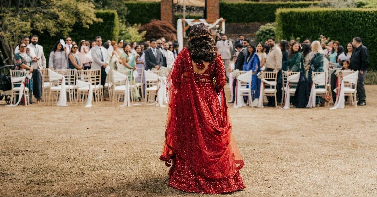 A woman in a red traditional dress walks away from a group of guests at an outdoor wedding ceremony.