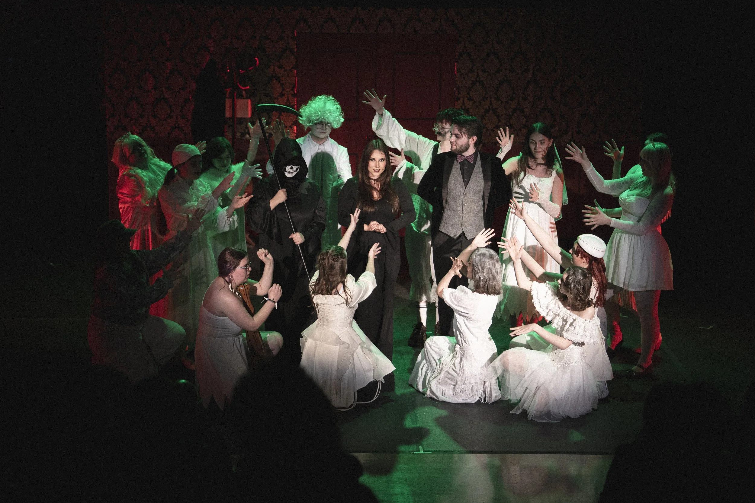 A theatrical scene with actors in period costumes, some kneeling and some standing, in a dimly lit room with dark, patterned wallpaper. The actors are engaged in a dramatic or musical performance.