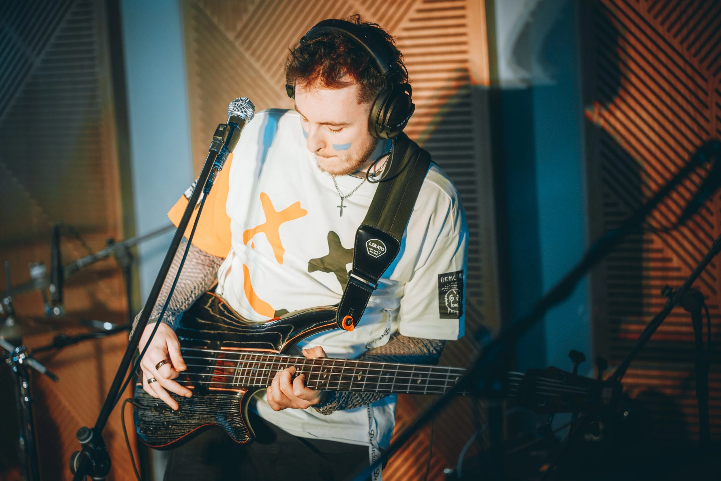 A young man with curly hair wearing headphones is performing in a music studio, playing a bass guitar and standing in front of a microphone.