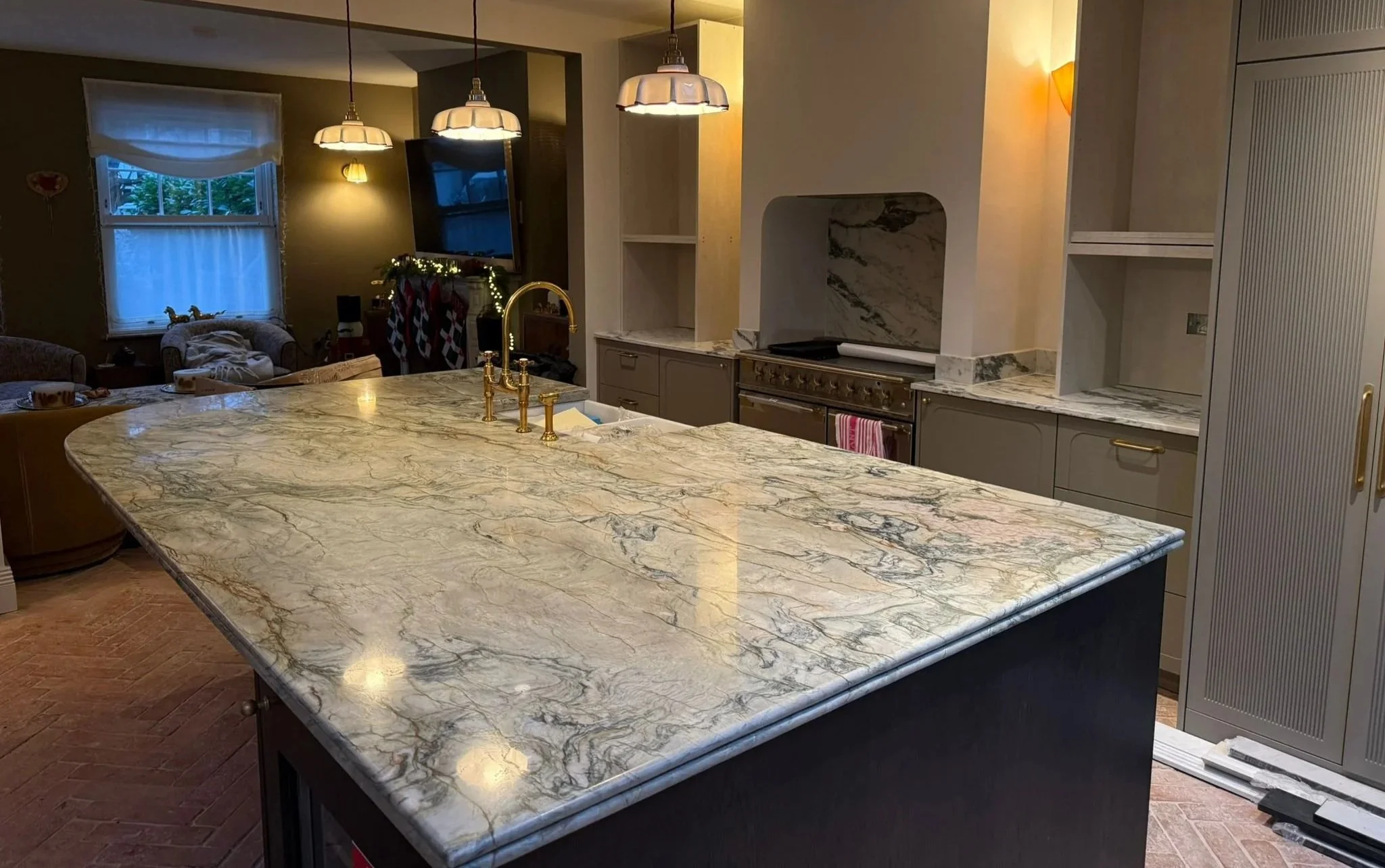 A large kitchen island with a polished marble countertop featuring a gold faucet and sink. In the background, there are hanging pendant lights, a television, and a window with blinds. The kitchen has gray cabinets and a stove with a marble backsplash.