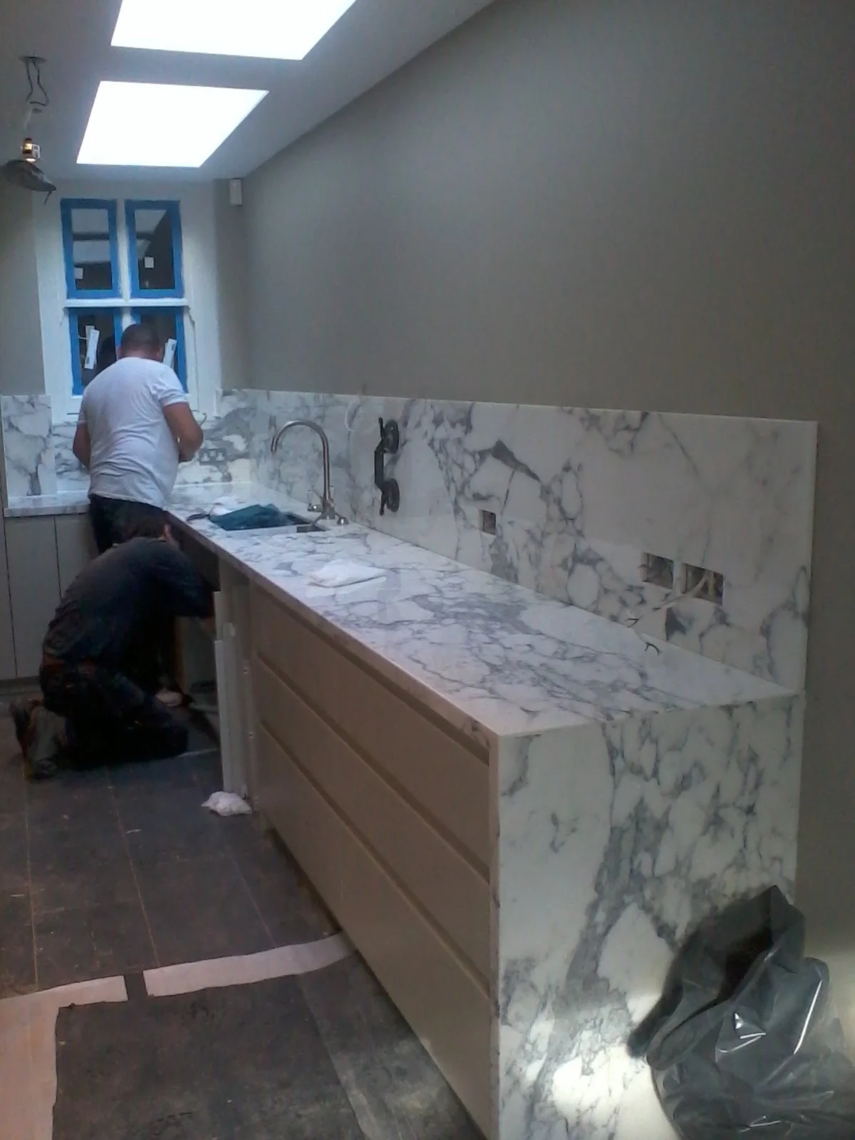 Two workers installing a marble countertop in a kitchen with gray walls and a window at the back.