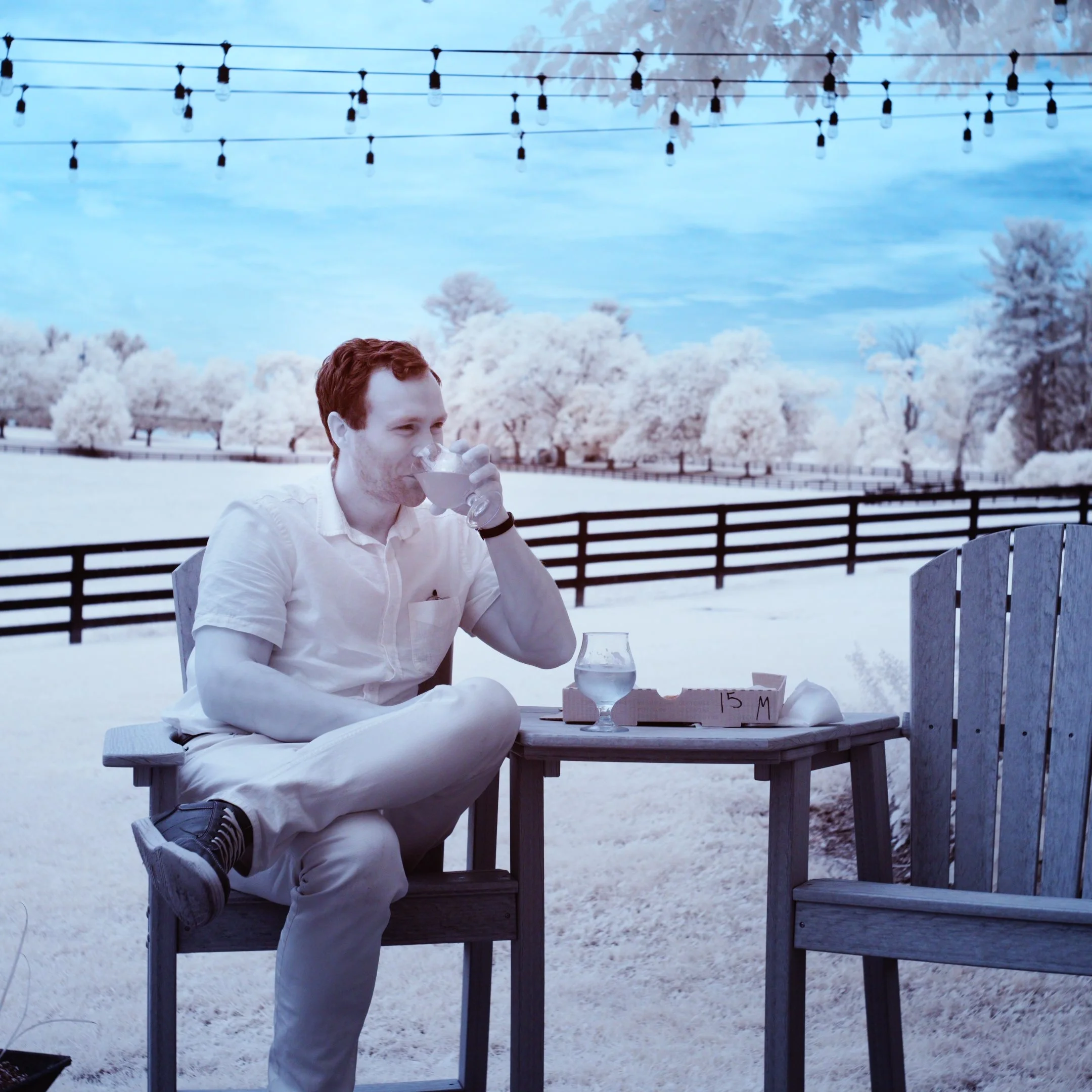 A person sits at an outdoor table enjoying a drink. Use of an infrared camera creates a surreal landscape of white folliage and blue sky on a summer day.