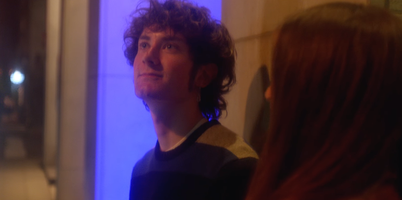 A man with curly hair smiling and looking upward, standing indoors against a blue-lit background, with a partially visible woman is visible in the foreground.