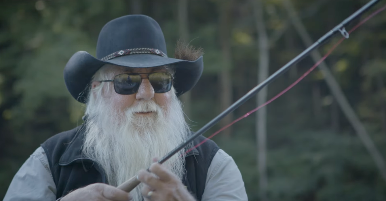 Older man with white beard wearing sunglasses, a black cowboy hat, and a vest, fishing with a rod in an outdoor natural setting.
