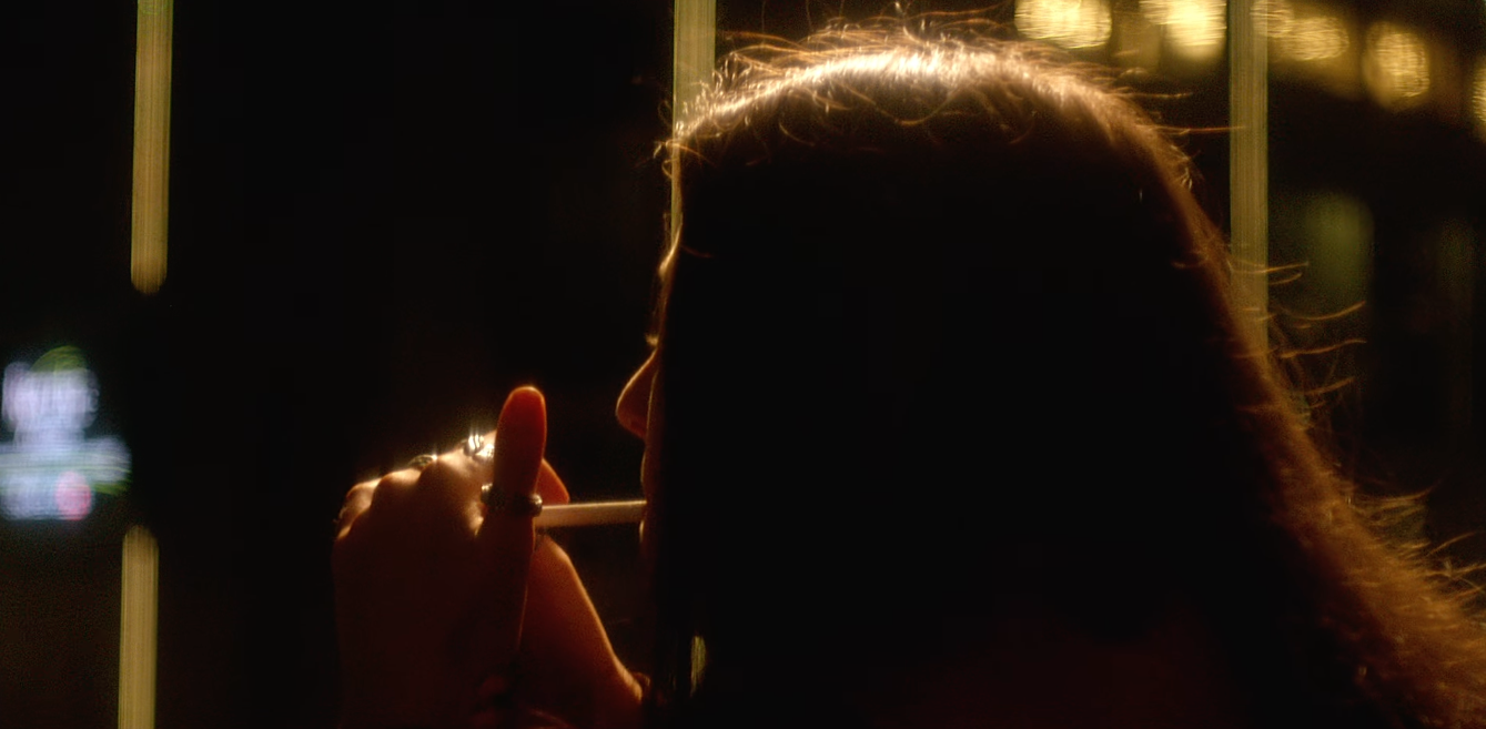 Silhouette of a person with long hair, holding a cigarette, in a dimly lit environment.