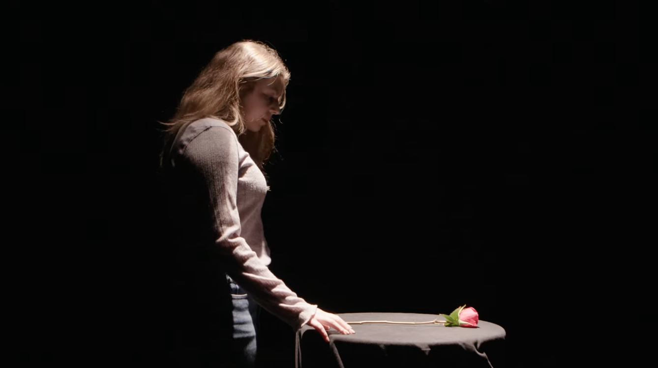 A woman with long blonde hair standing next to a black table with a pink rose on it, in a dark setting.