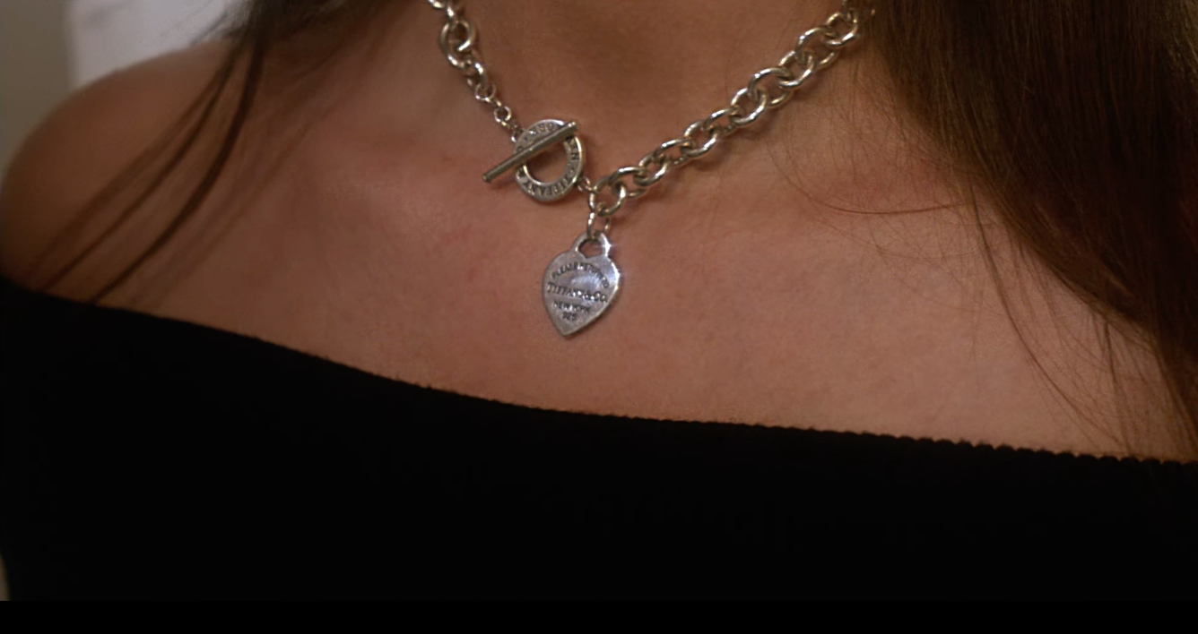 Close-up of a person's neck and chest wearing two layered silver necklaces, one with a heart-shaped pendant and the other with a toggle clasp.