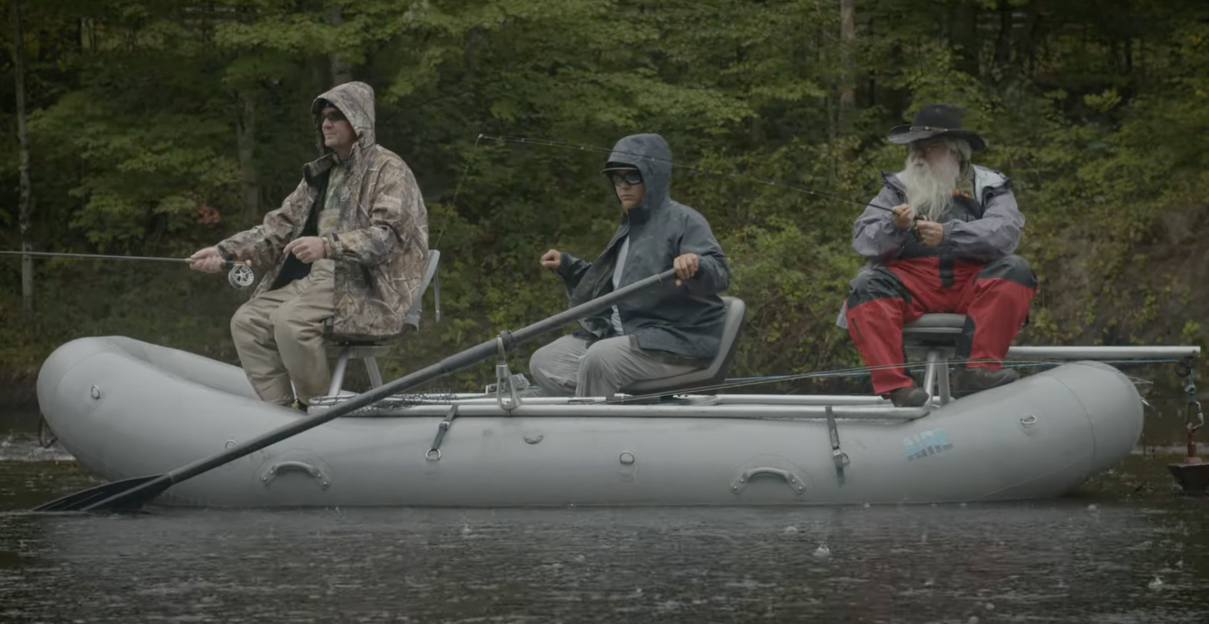 Three people fishing from an inflatable boat on a lake with green trees in the background.