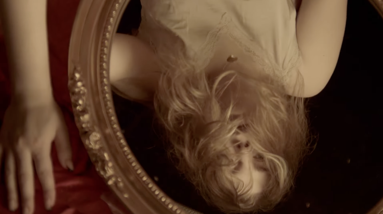 A person with long, curly blonde hair taking a selfie in a mirror, wearing a light-colored dress, with their head tilted back. The mirror has a decorative border. Part of their hand is visible in the lower left side of the image.
