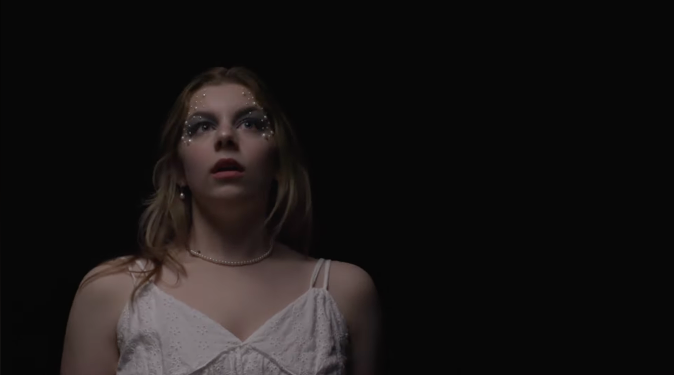 A woman with light skin and long blonde hair looking upward, wearing a white lace top, a pearl necklace, and makeup with decorative rhinestones near her eyes, set against a black background.