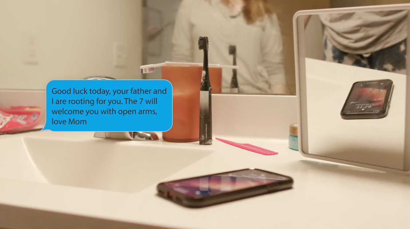 A bathroom countertop with a smartphone, toothbrush in a cup, a small container, and a mirror reflecting a person. Blue text bubble message saying, 'Good luck today, your father and I are rooting for you. The 7 will welcome you with open arms, love M
