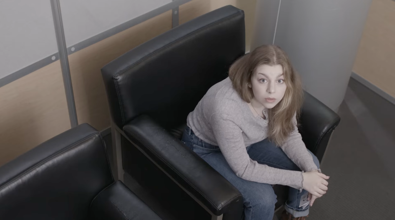 A young woman with long, wavy blonde hair sitting on a black leather bench in what appears to be a waiting area, looking up towards the camera with a curious expression.