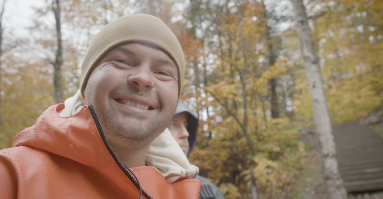 Close-up of a smiling man in an orange rain jacket and beige beanie outdoors in a forest with autumn foliage.