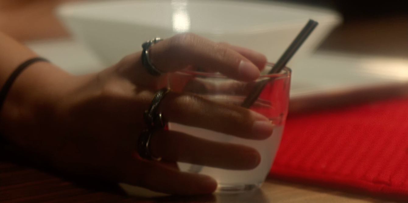 A person's hand holding a glass of water with a metal straw, resting on a wooden table with a red placemat.