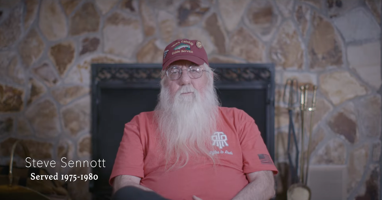 An elderly man with a long white beard and glasses sits in front of a stone fireplace. He is wearing a maroon cap and a red T-shirt with a logo. Text on the image indicates his name is Steve Sennott and he served from 1975 to 1980.