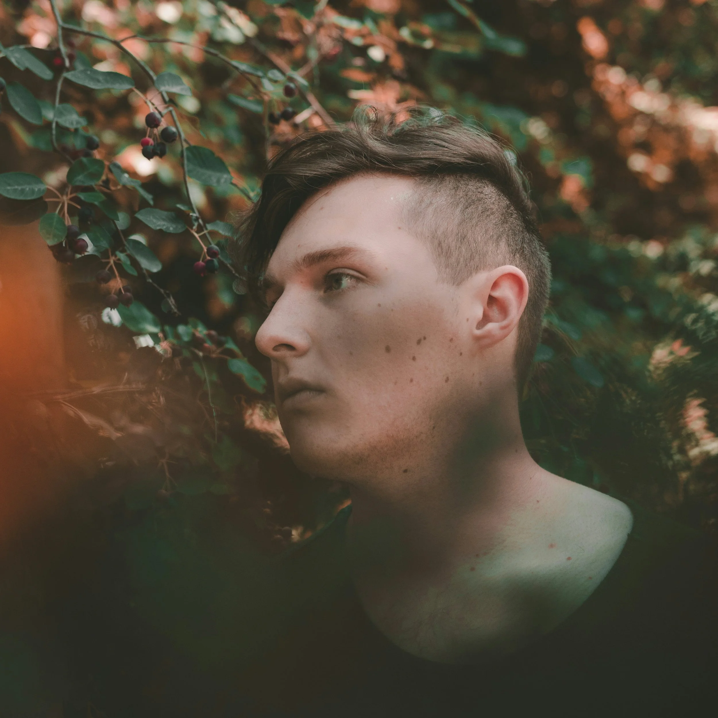 A person's face partially visible, with short hair, green eyes, and freckles, surrounded by green leaves and small black berries on a tree branch in a natural outdoor setting.