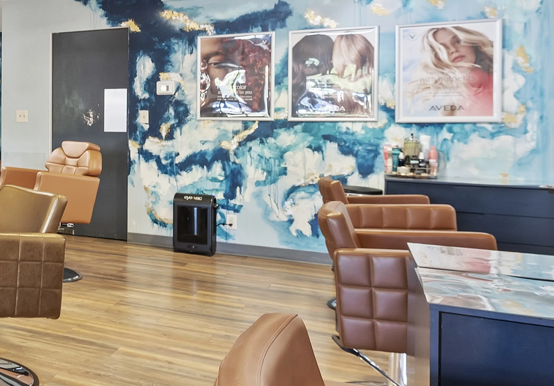 An empty waiting or lounge area with a colorful abstract mural on the wall. Several brown leather chairs and tables are arranged around the room, with some items on a counter and an air purifier in the foreground.