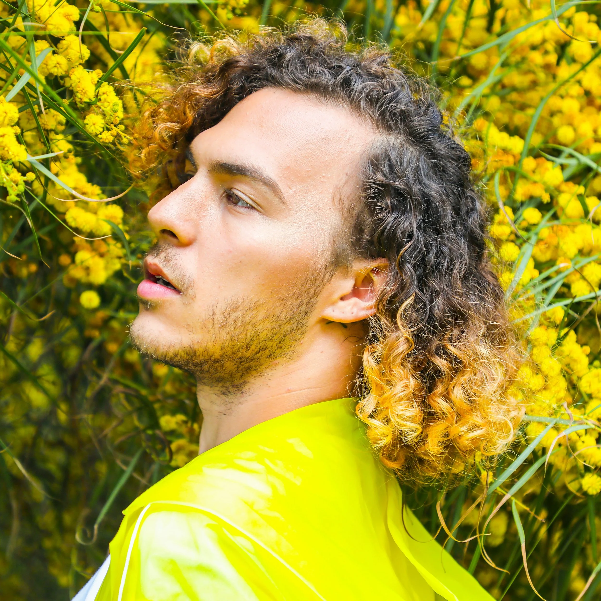 Close-up of a person's face with curly hair, partially visible, surrounded by yellow flowers and green foliage.